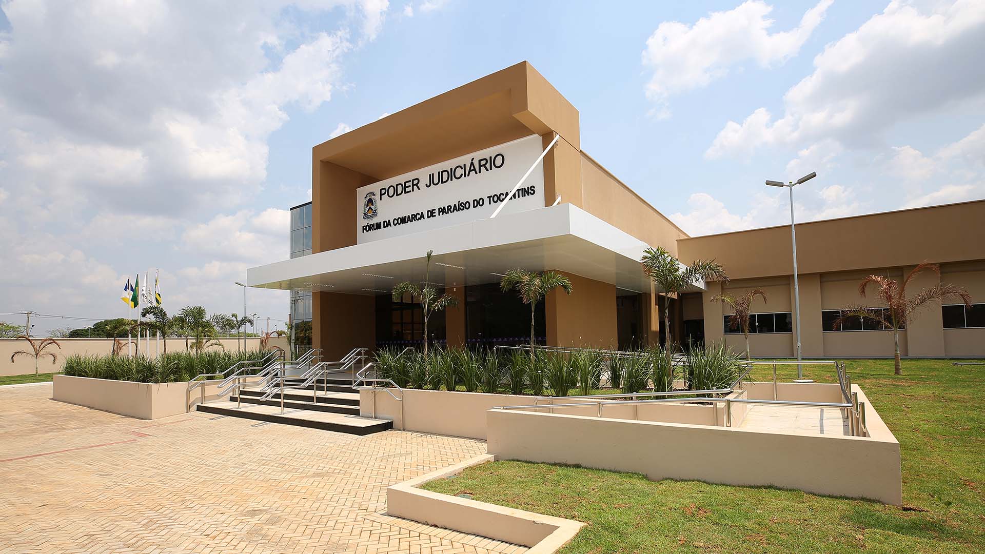 Prédio nas cores marrom queimado, branco e na esquerda uma parede espelhada, a fachada tem uma escada com dois corrimão e ao lado das escadas umas plantas pequenas bem verdes