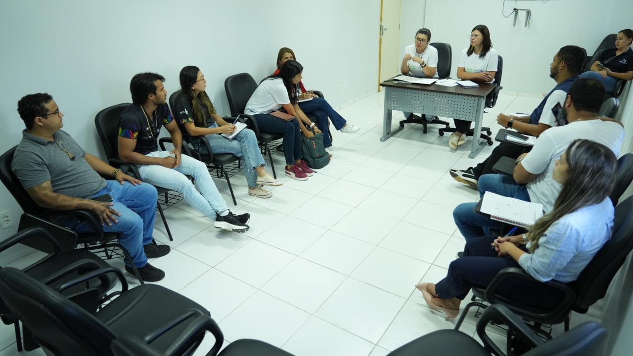 A imagem mostra uma sala branca, pessoas estão sentadas em círculos em cadeiras pretas, entre elas estão homens e mulheres, como se estivessem em reunião, todas elas estão olhando para duas mulheres de camiseta branca sentadas atrás de uma mesa e elas estão falando, a mulher da esquerda é branca, usa óculos e estar com o cabelo preso para trás, a mulher da direita é branca, tem o cabelo preto na altura dos ombros e usa uma sandália de cor branca