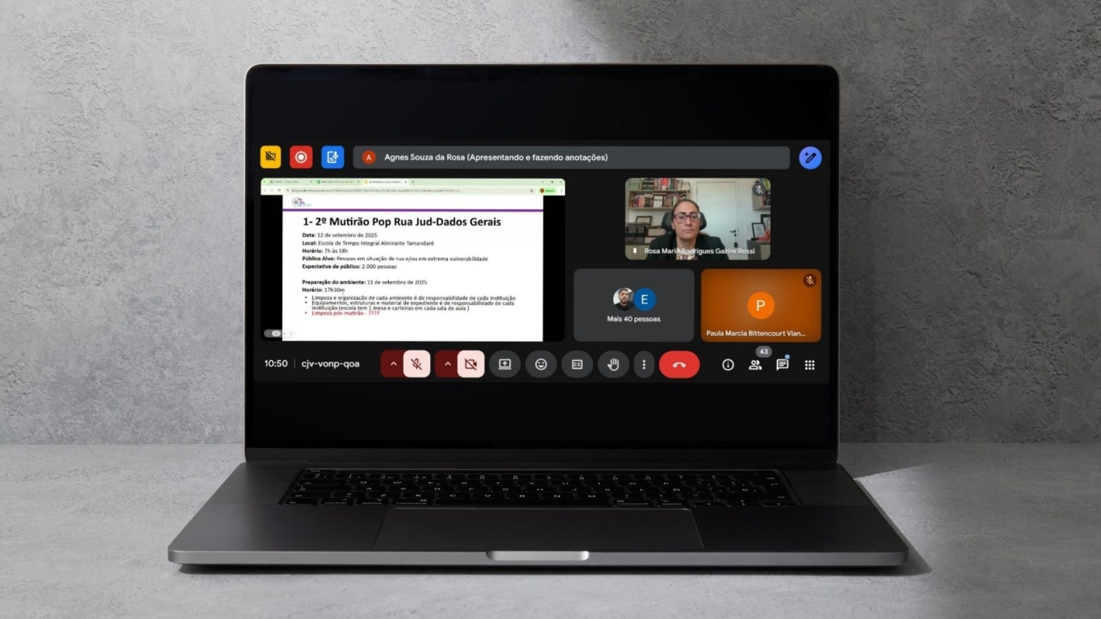 A imagem mostra um laptop com uma videochamada acontecendo. Na tela, uma mulher está apresentando um slide sobre um evento chamado "Mutirão Pop Rua", que será em setembro de 2025 para ajudar pessoas em situação de rua. Também aparecem várias janelas com outras pessoas participando da reunião. A interface mostra controles típicos de videochamada, como microfone e câmera. O laptop está sobre uma superfície cinza.