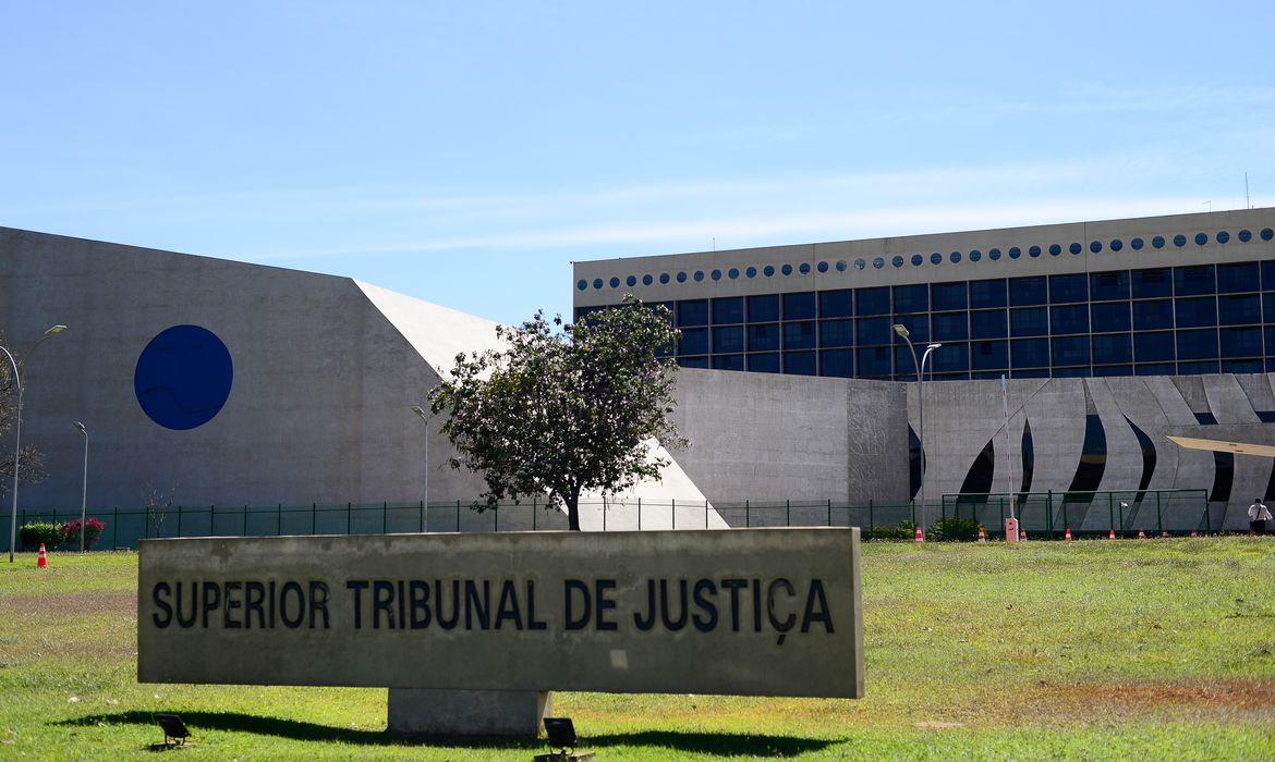 Vista do edifício do STJ. O prédio é moderno, com estrutura cinza claro, com grandes janelas e um design angular e simétrico. À frente uma placa indicando "Superior Tribunal de Justiça" e uma área de grama verde.
