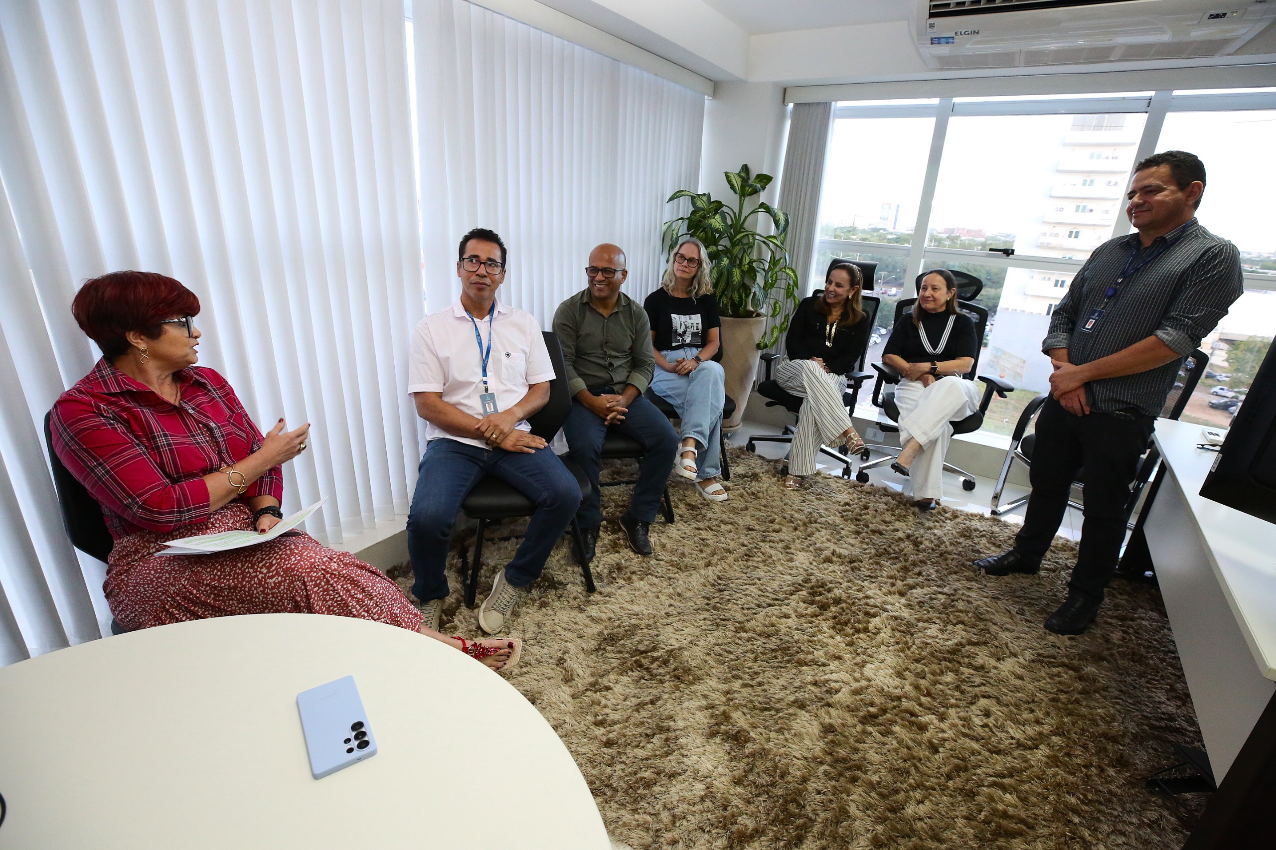 A imagem mostra um grupo de pessoas em uma sala de escritório, sentadas em cadeiras ao redor de uma mesa redonda. Há sete pessoas, sendo seis mulheres e um homem, todas vestindo roupas casuais de trabalho, como camisas e blazers. Elas parecem estar participando de uma reunião, com expressões atentas e concentradas. A sala é bem iluminada, com uma grande janela ao fundo que oferece uma vista da cidade. O ambiente é moderno e acolhedor, com um carpete claro no chão. Na mesa, há um celular pequeno, mas não há muitos outros objetos visíveis.