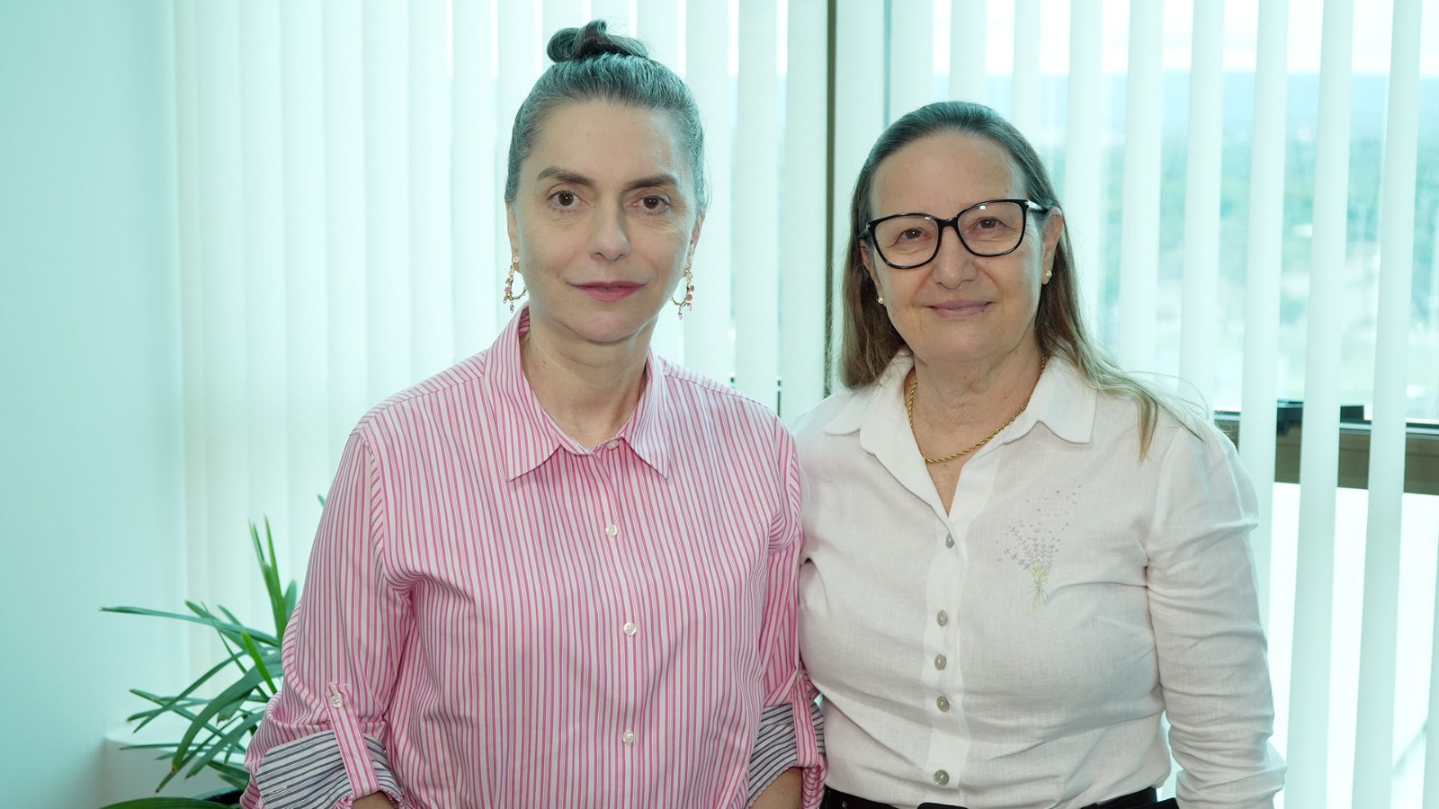 Desembargadora Maysa, à direita, de blusa branca, e a conselheira Doris Coutinho, de blusa rosa, posam para foto em um ambiente interno.