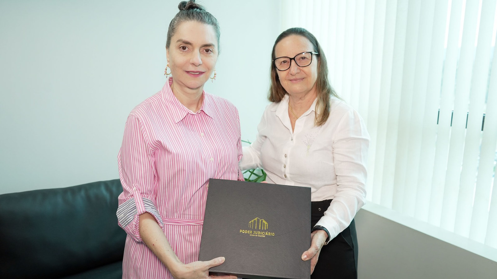 Desembargadora Maysa, à direita, de blusa branca, entrega uma caixa marrom com o nome Poder Judiciário na capa à conselheira Doris Coutinho, de blusa rosa.
