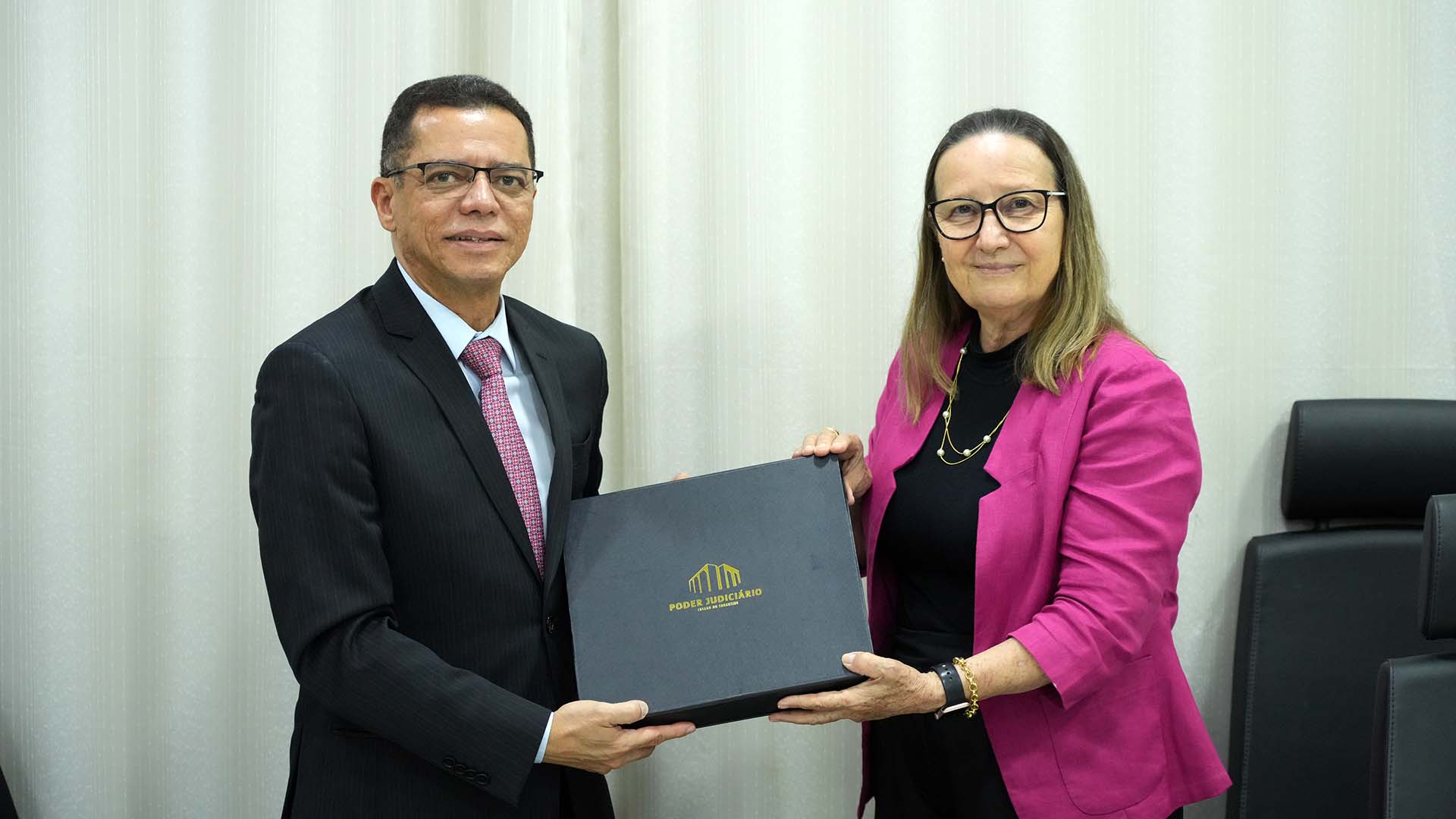 Desembargadora Maysa, à direita,  de roupa preta e blazer rosa, entrega uma caixa marrom com a logo do Poder Judiciário ao desembargador do TRT, de terno preto, camisa branca e gravata vermelha.  