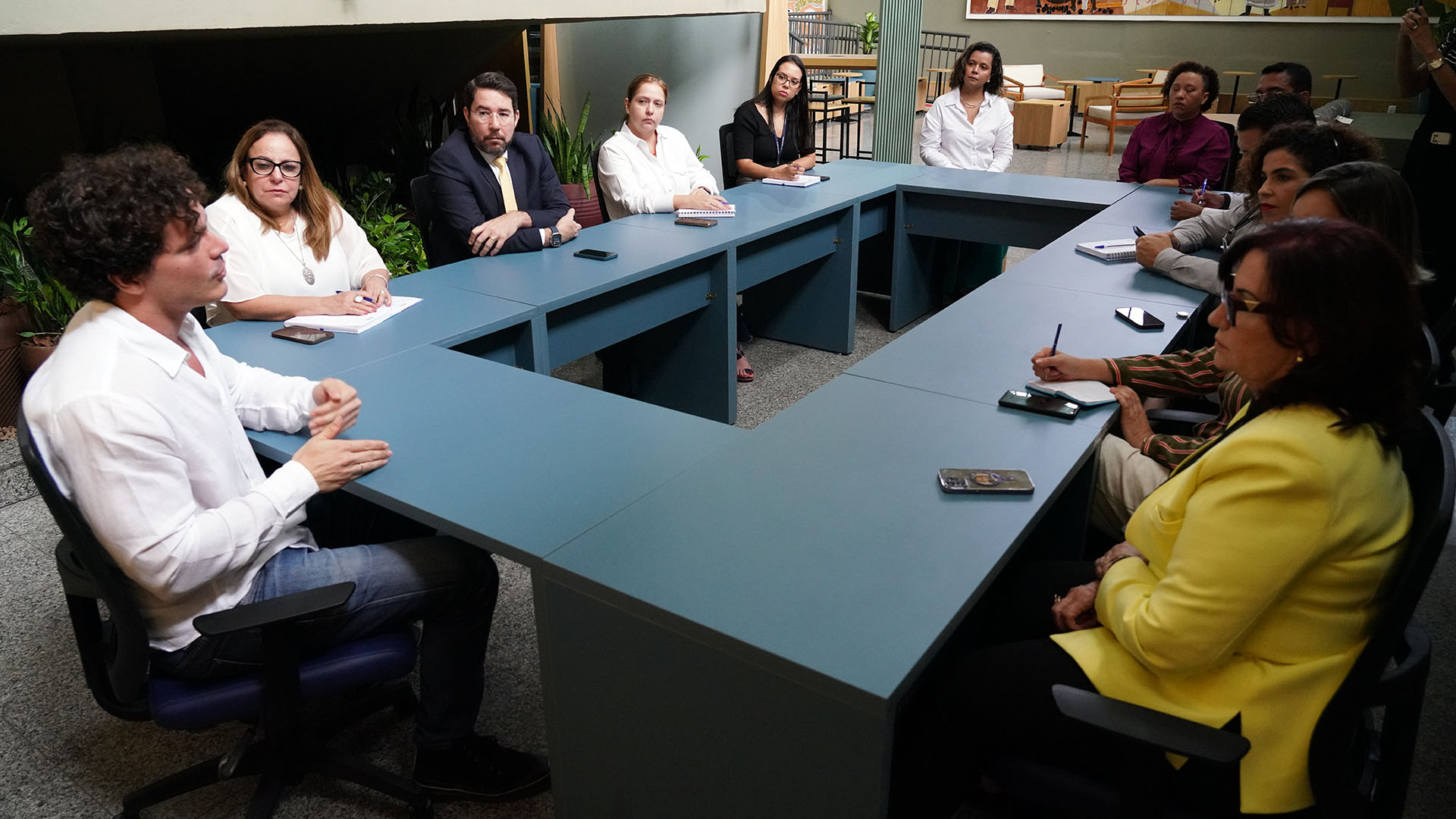 Pessoas sentada a mesa durante reunião, um homem de blusa branca social fala enquanto as outras pessoas escutam