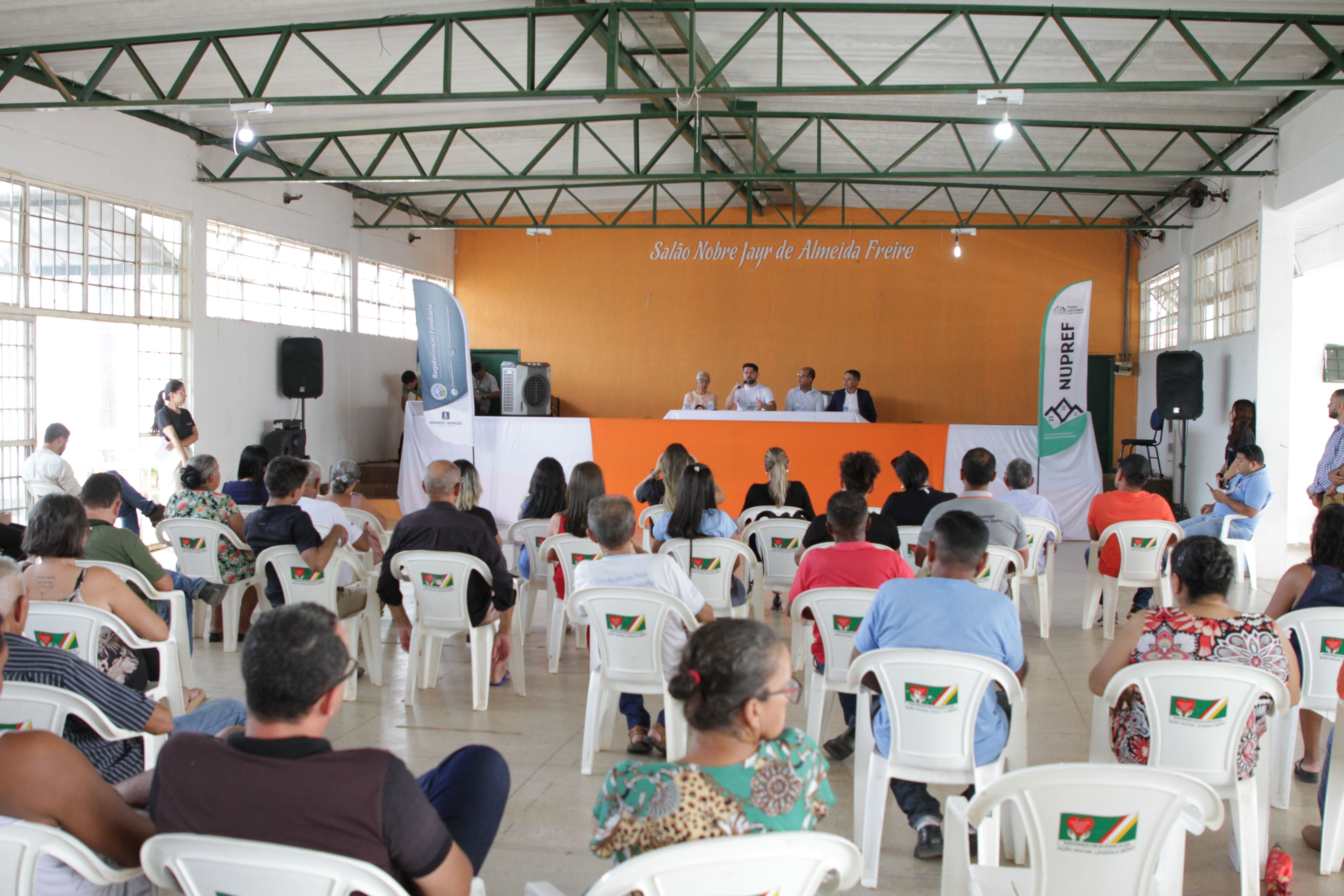 Pessoas sentadas em frente à um palco assistindo audiência pública 