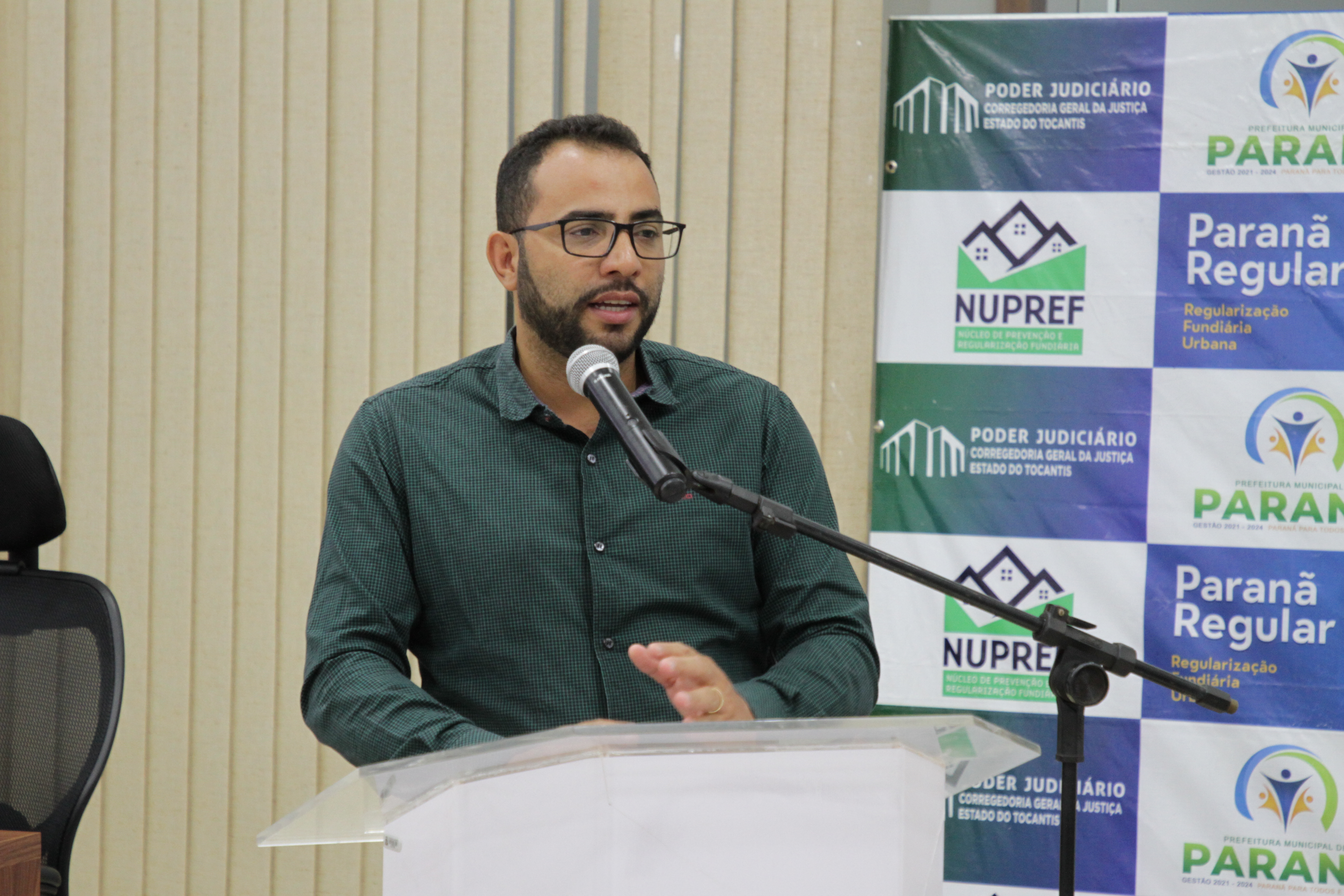 Homem de cabelo preto com blusa social verde escura fala ao microfone