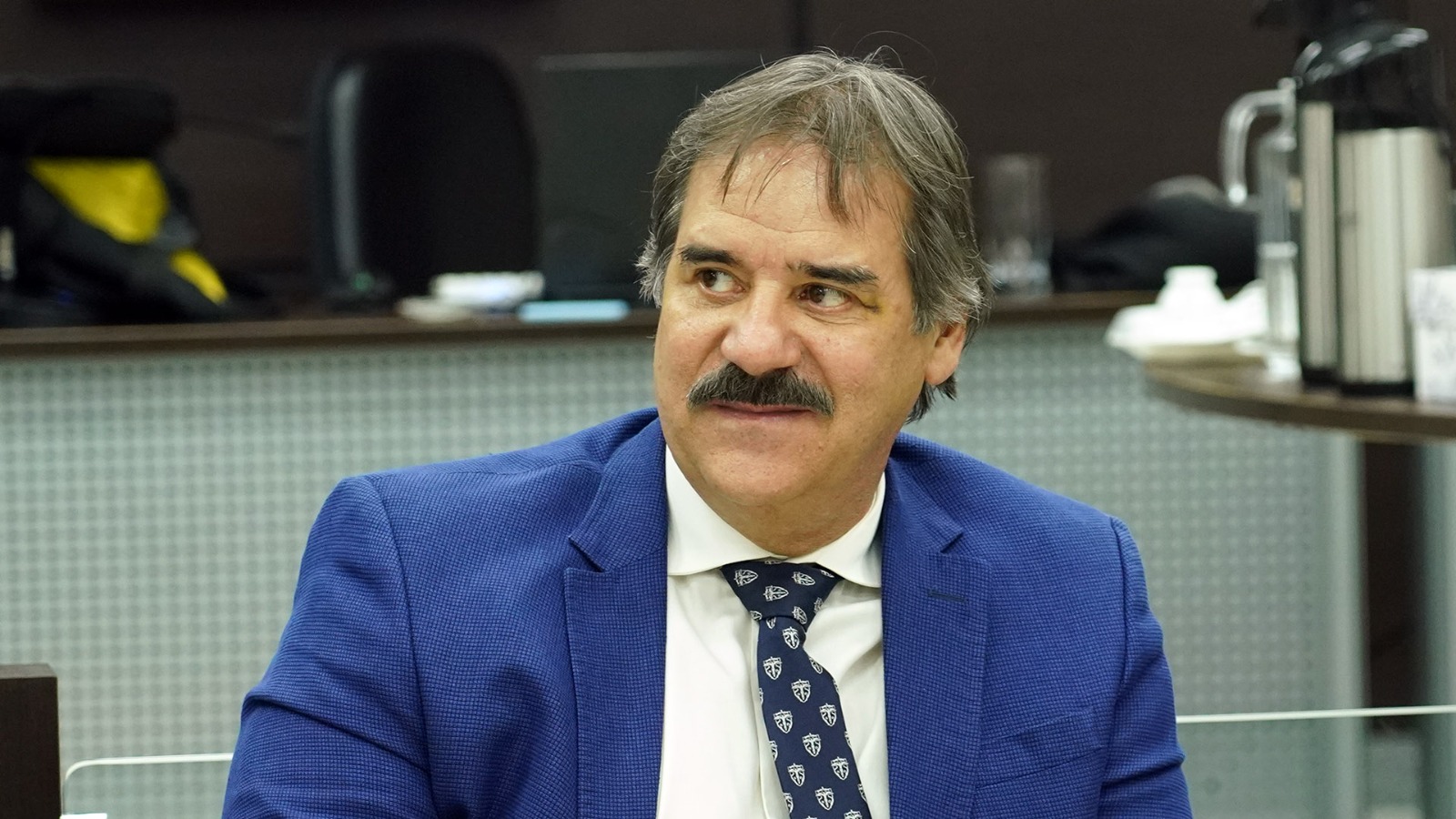 Homem de terno azul marinho, camisa branca e gravata azul escura, cabelo preto com alguns fios brancos, ele tem bigode e estar olhando para o lado esquerdo