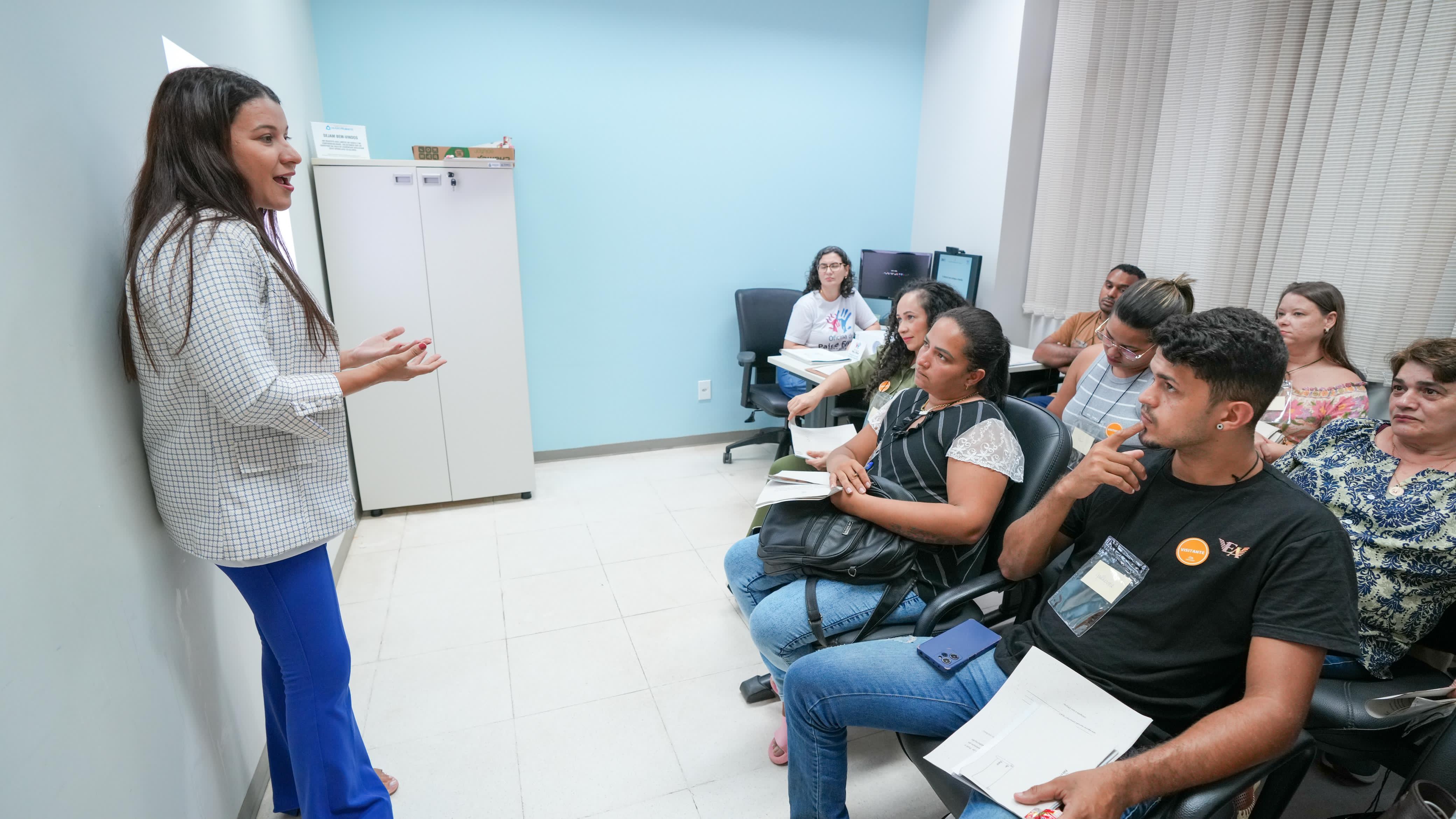 Sala na cor azul claro, mulher em pé do lado esquerdo, com um blazer xadrez e uma calça azul, o cabelo dela é preto e estar solto, ela gesticula com as mão como se estivesse conversando, ao fundo tem um armário branco, de frente para mulher do lado direito tem um grupo de pessoas, homens e mulheres que estão olhando pra ele.