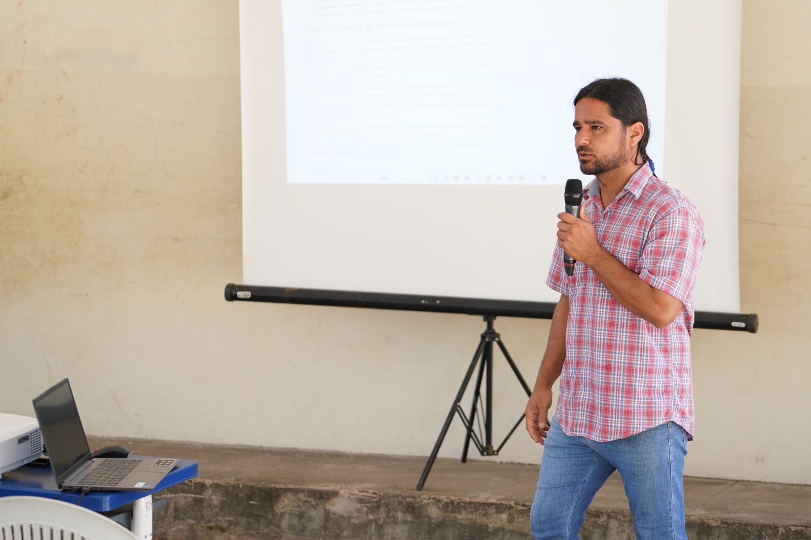 Agrônomo, um homem moreno com barba curta e cabelo grande amarrado, usando camisa quadriculada em tons de vermelho e azul e calsa jeans
