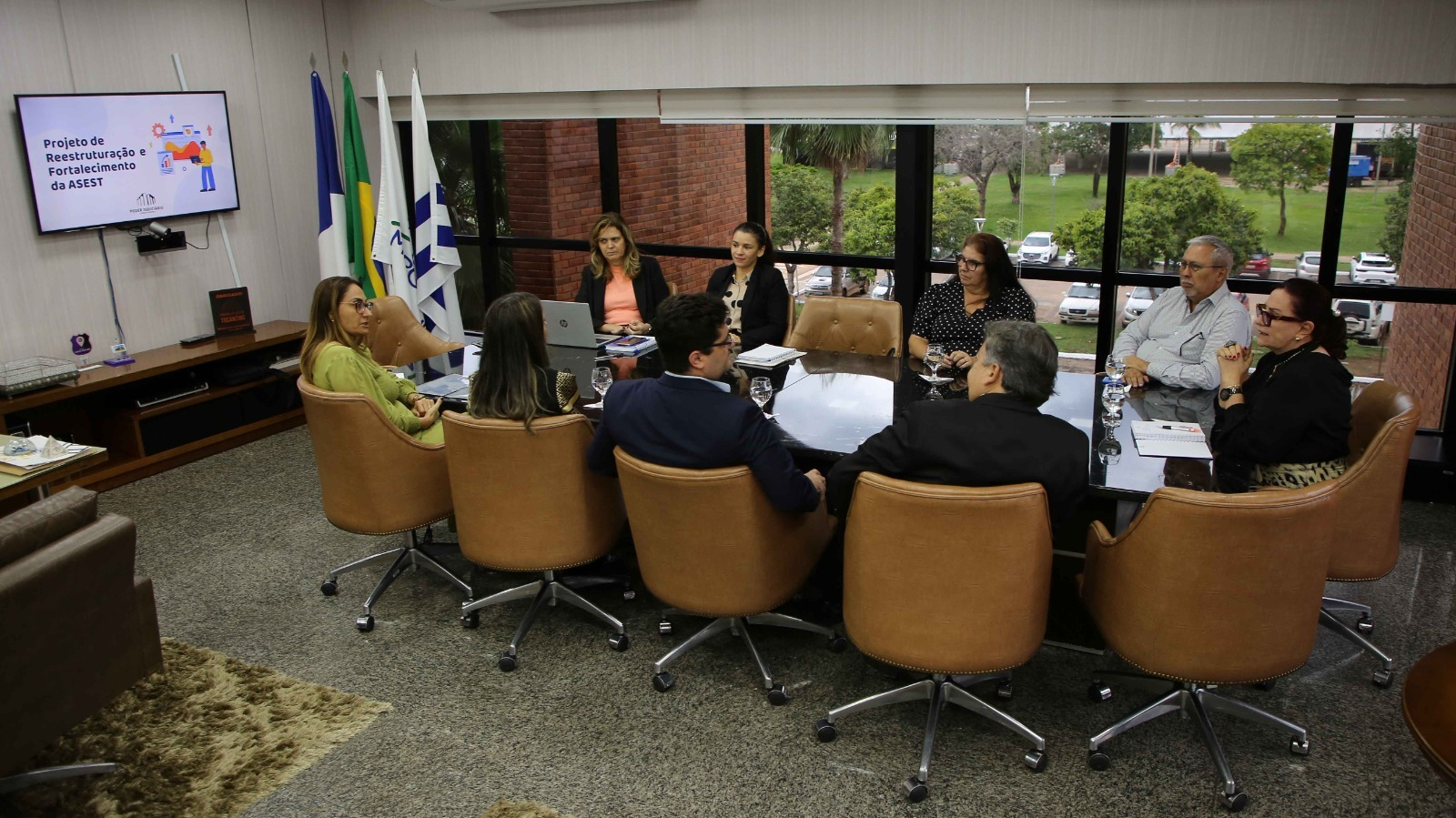 Imagem mostra pessoas sentadas em torno da mesa na presidência do TJTO durante apresentação de projeto em televisor na pared