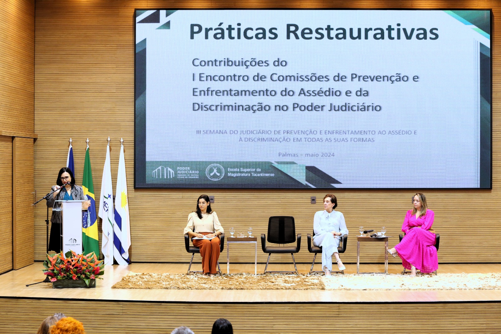 Pauco do auditório onde estão a palestrante, no púlpito, falando ao microfone, à esquerda e ao centro a desembargadora Angela Haonat, entre as juízas Julianne (d) e Suzidarly (e), todas sentadas assistendo a palestra
