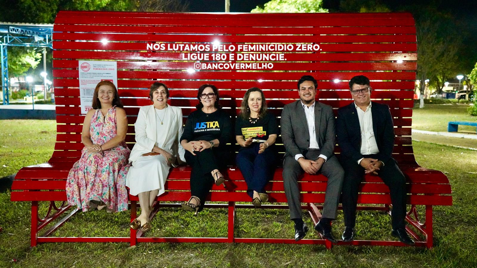 Um banco enorme de cor vermelha e nele estão sentadas cinco pessoas, quatro mulheres e dois homens, todos de cabelo preto, os homens estão de terno, calça e sapato social e as mulheres estão uma de vestido colorido, a outra vestido branco, a do lado calça preta e blusa preta e a do meio também