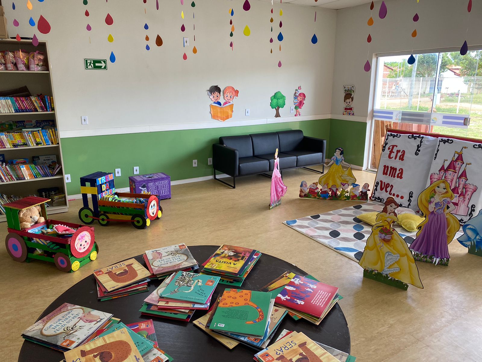 Foto de uma biblioteca infantil com parede branca e verde chão em madeira com uma mesa redonda com livros empilhados uma estantes com mais livros um banco preto ao lado da estante e dois berços pequenos a frente da estante com mais livros