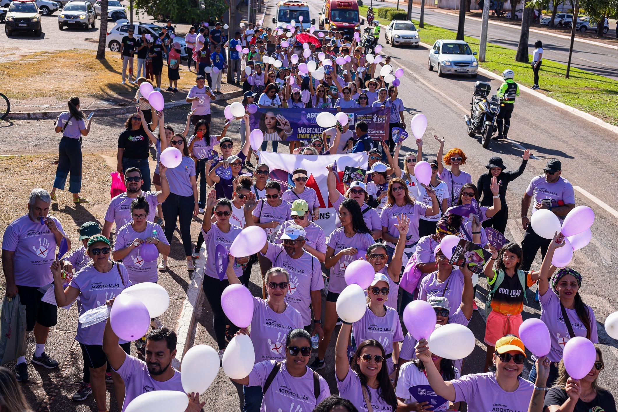 Várias pessoas vestidas com camiseta lilás, com balões brancos e lilás, e com faixas e cartazes participam de uma caminhada de combate à violência contra a mulher em uma via púbica