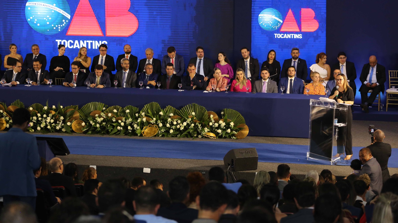 Palco com uma grande mesa coberta com uma toalha azul, várias pessoas sentadas, vestindo trajes sociais.