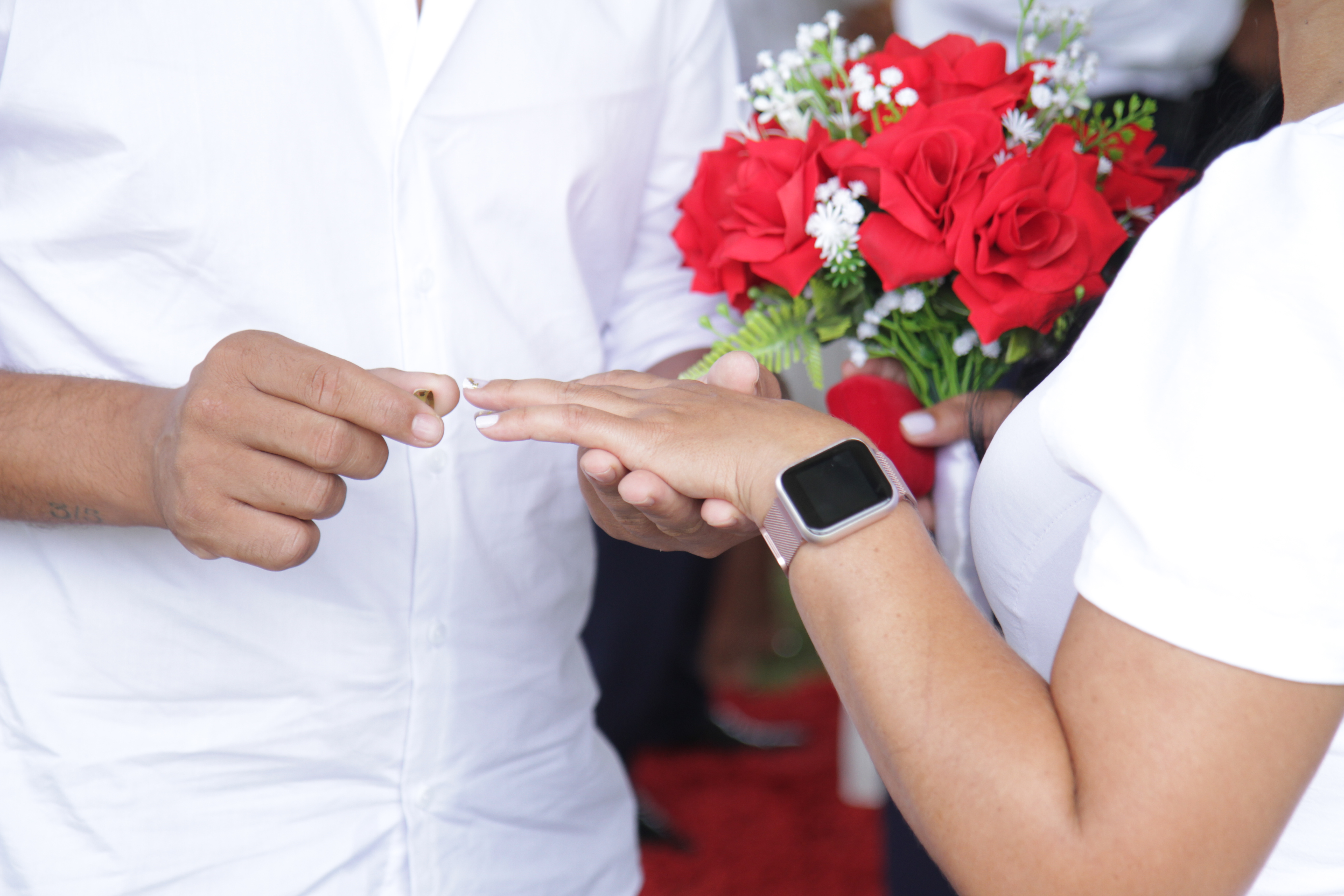 Imagem de um casamento focada na mão do casal trocando alianças e a noiva segura um buquê de flores vermelhas.