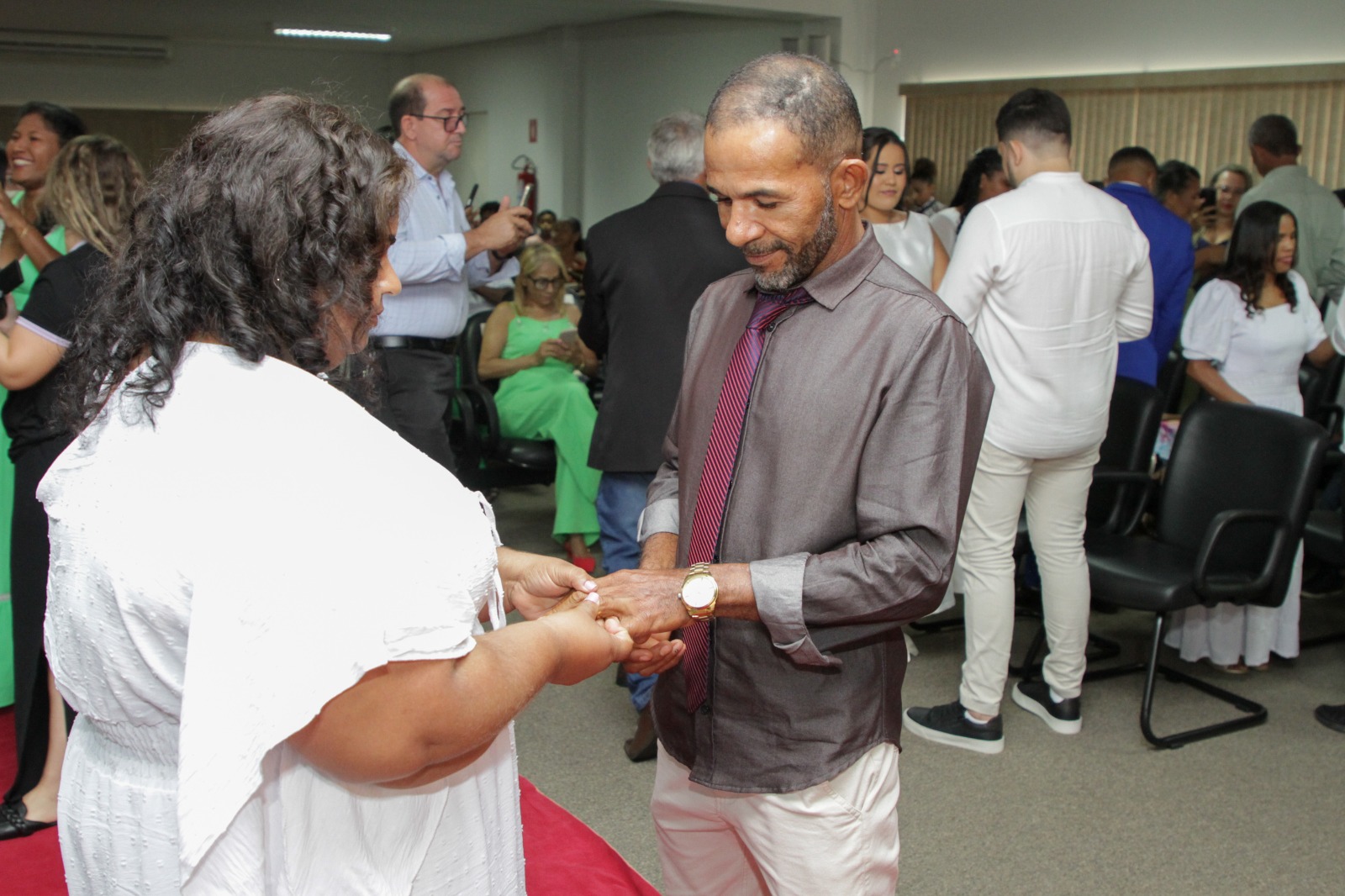 Mulher de vestido branco e cabelo preto coloca anel no dedo de um homem com camisa social preta e gravata vermelha e calça bege