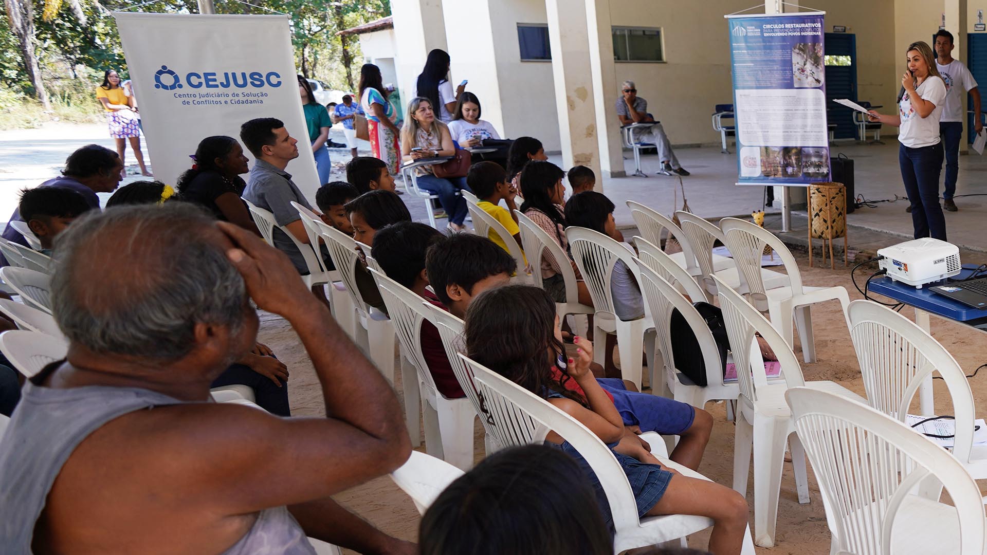 Vários indígenas Krahô sentados em cadeiras branças assistindo a palestra de uma facilitadora, uma jovem loura em pé, seghurando um microfone, vestida com uma camiseta branca com alogo da Justiça Restaurativa e Calça Jeans 