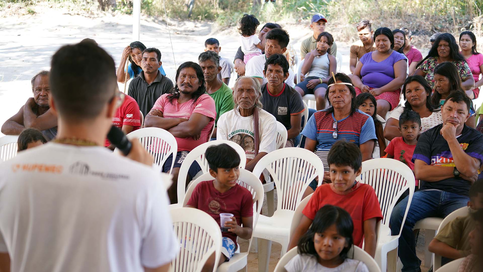 Vários indígenas krahô sentados em cadeiras brancas de frente ouvindo um facilitador do projeto, que está de costa, usando camiseta branca.