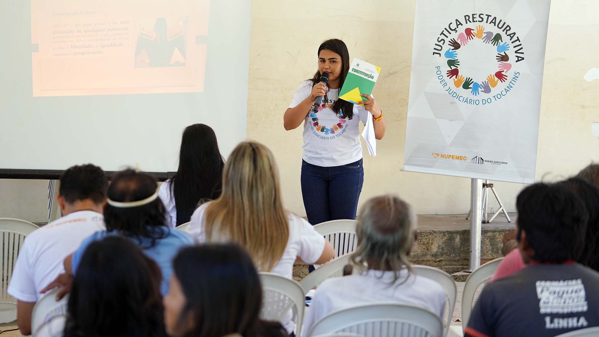 Facilitadora em pé, de frente, com um exemplar da Cosntituição em mãos, falando aos indígenas Krahô, que está de costa. Ao lado da facilitadora, um banner com a logo do Projeto Justiça Restaurativa