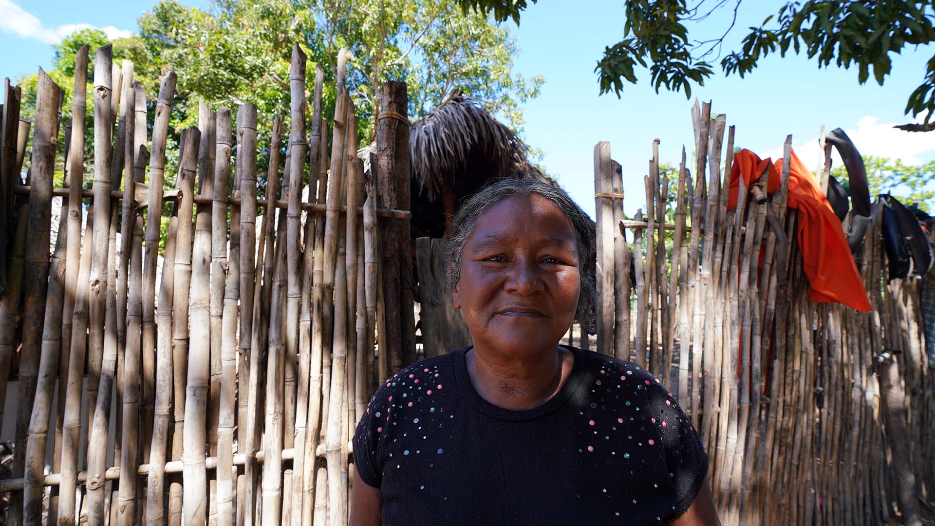 Cleonice Krahô, uma senhora que usa blusa preta com detalhes coloridos e cabelos grisalhos. Ao fundo uma cerca com estacas de madeira.