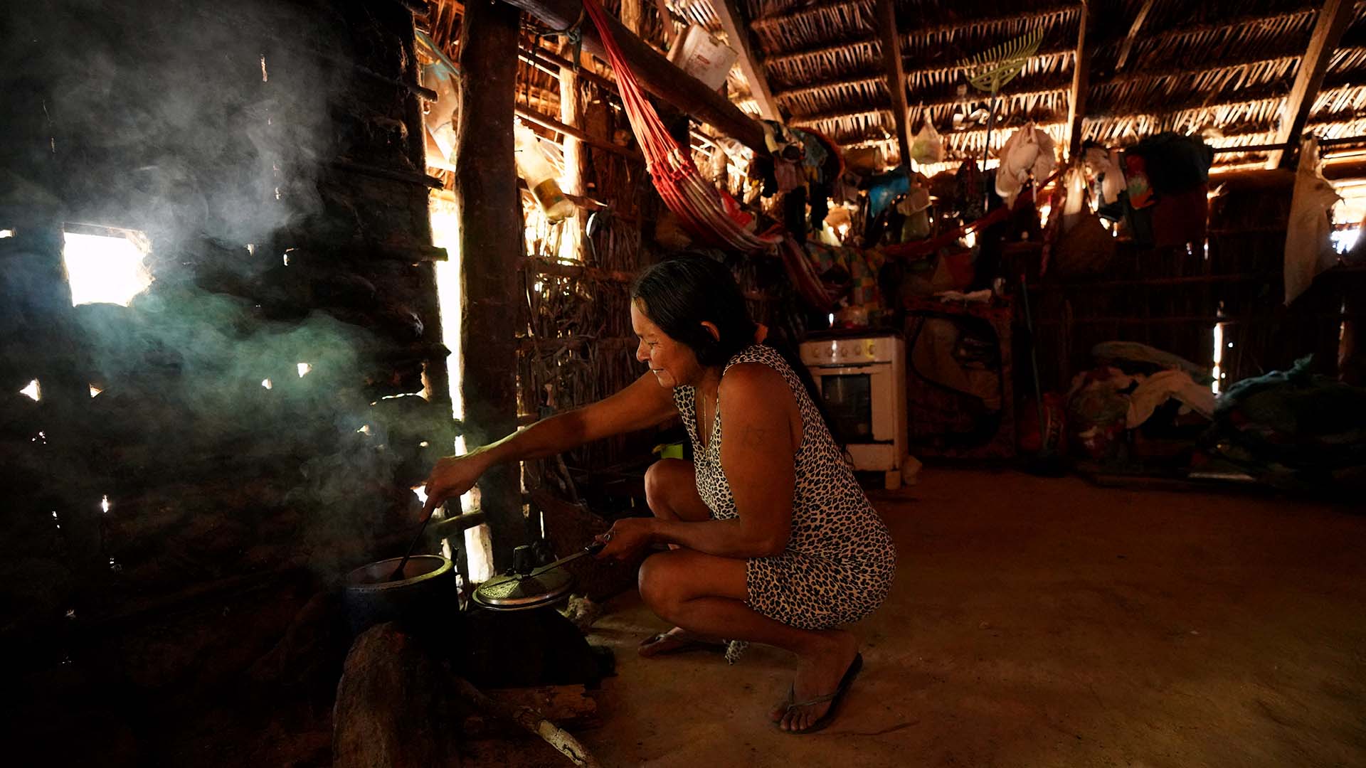 Rosilda Krahô, usando um vestido estampado em tons de preto e branco, agachada, mexendo a comida em uma panela preta que está em um fogo de lenhã no chão da sala da casa simples, coberta de palha e paredes de taipa e palha. Ao fundo um fogão.