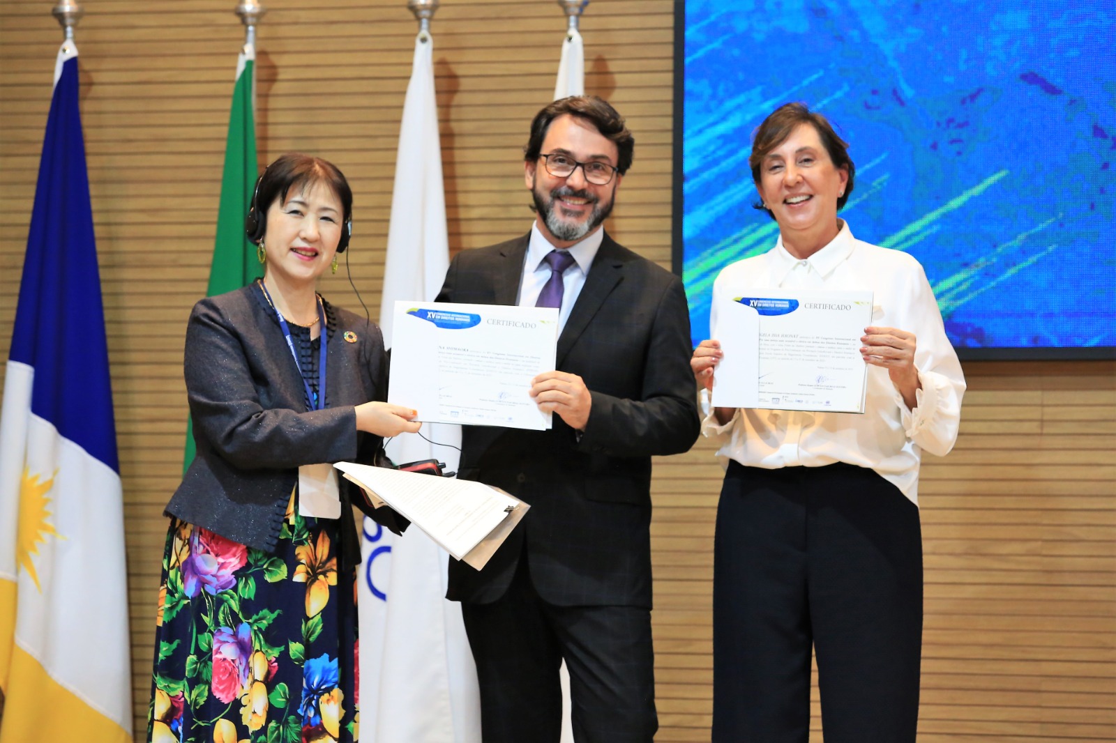 Mana Shimaoka, japonesa, de terno escuro e saia estampada, o professor Gustavo Paschoal ao centro, de terno e gravata escuros e camisa branca; e a desembargadora Angela Haonat à direita, de blusa branca e calça preta, segurando certificados
