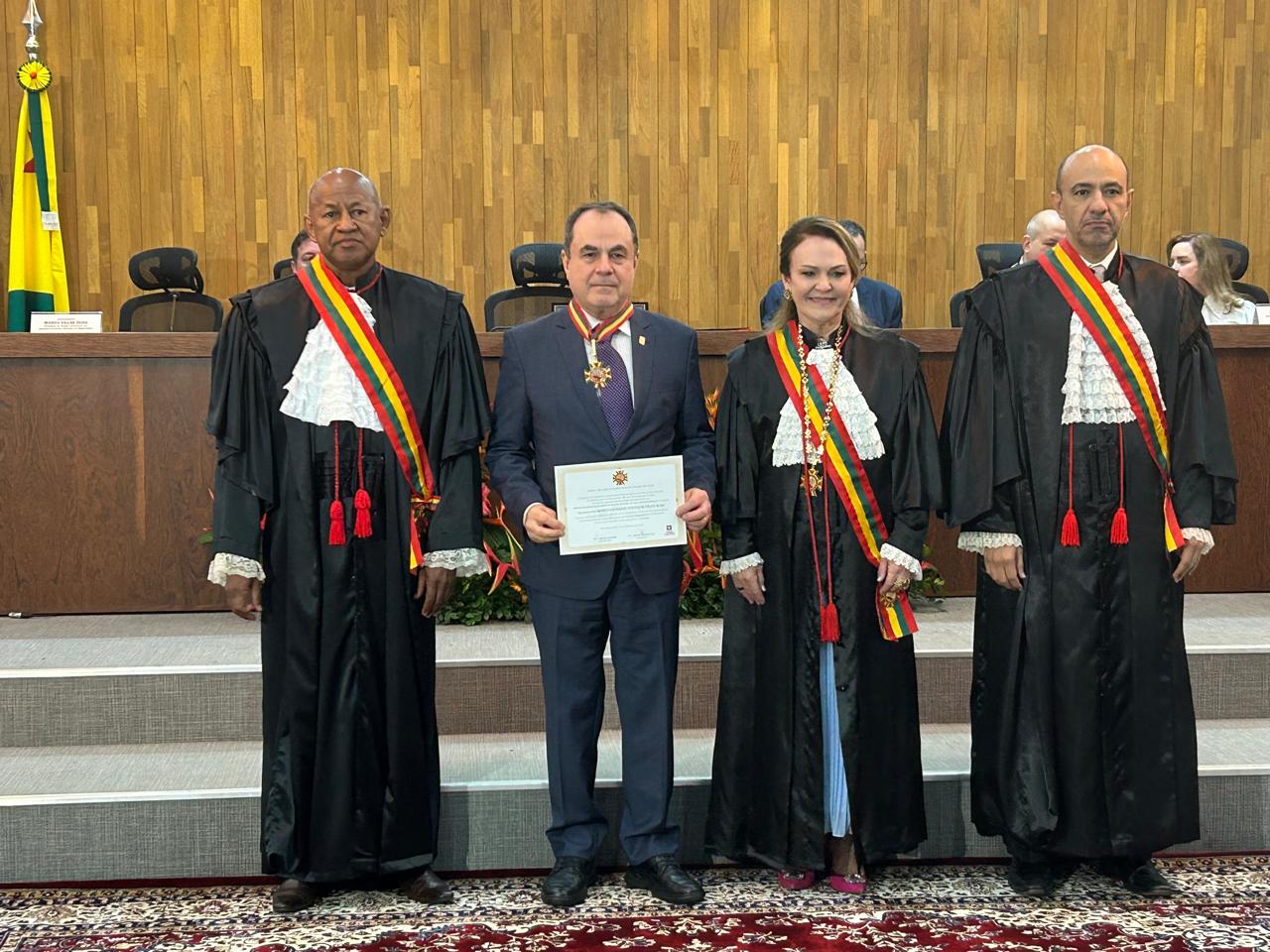 Quatros pessoas estão de pé, sendo três homens e uma mulher. Um dos homens segura nas mãos um laureado com o Mérito Judiciário.