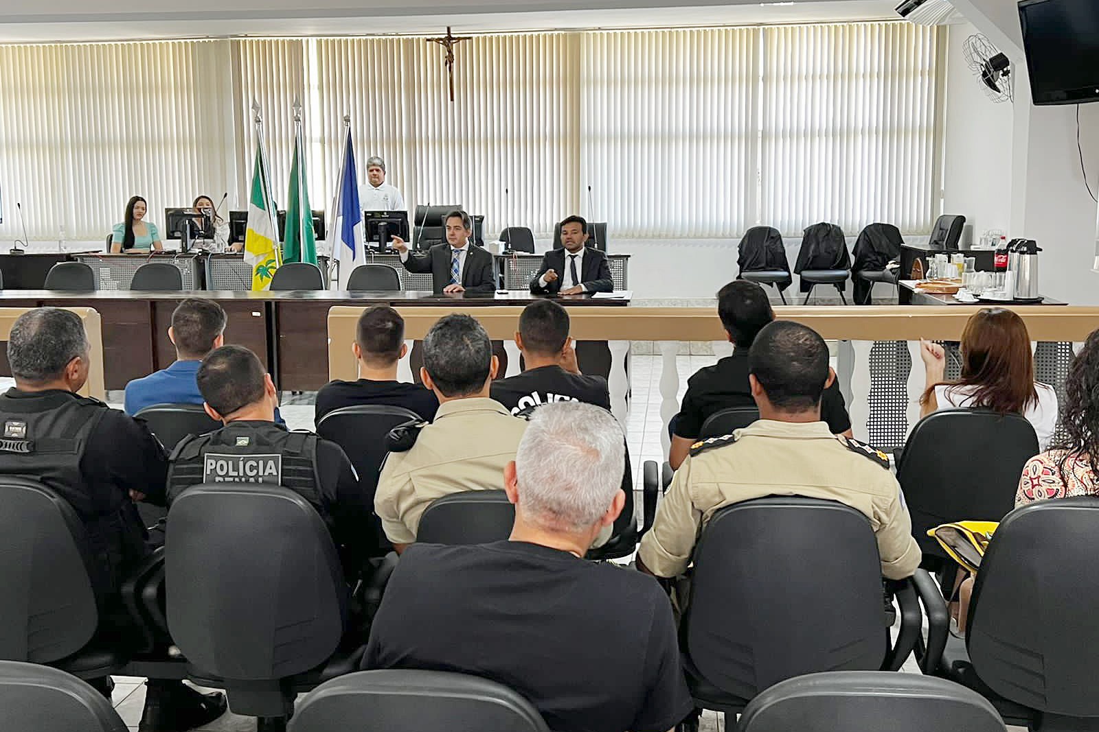 Imagem dw uma sala  com o juiz Helder Lisboa, um homem moreno e de terno e gravata preta e camisa branca, e o promotor Saulo Vinhal, um homem branco de cabelos grisalhos, terno escuro, camisa branca e gravata xadrez em azul e branco, sentados de frente e os policiais fardados sentados de costas 