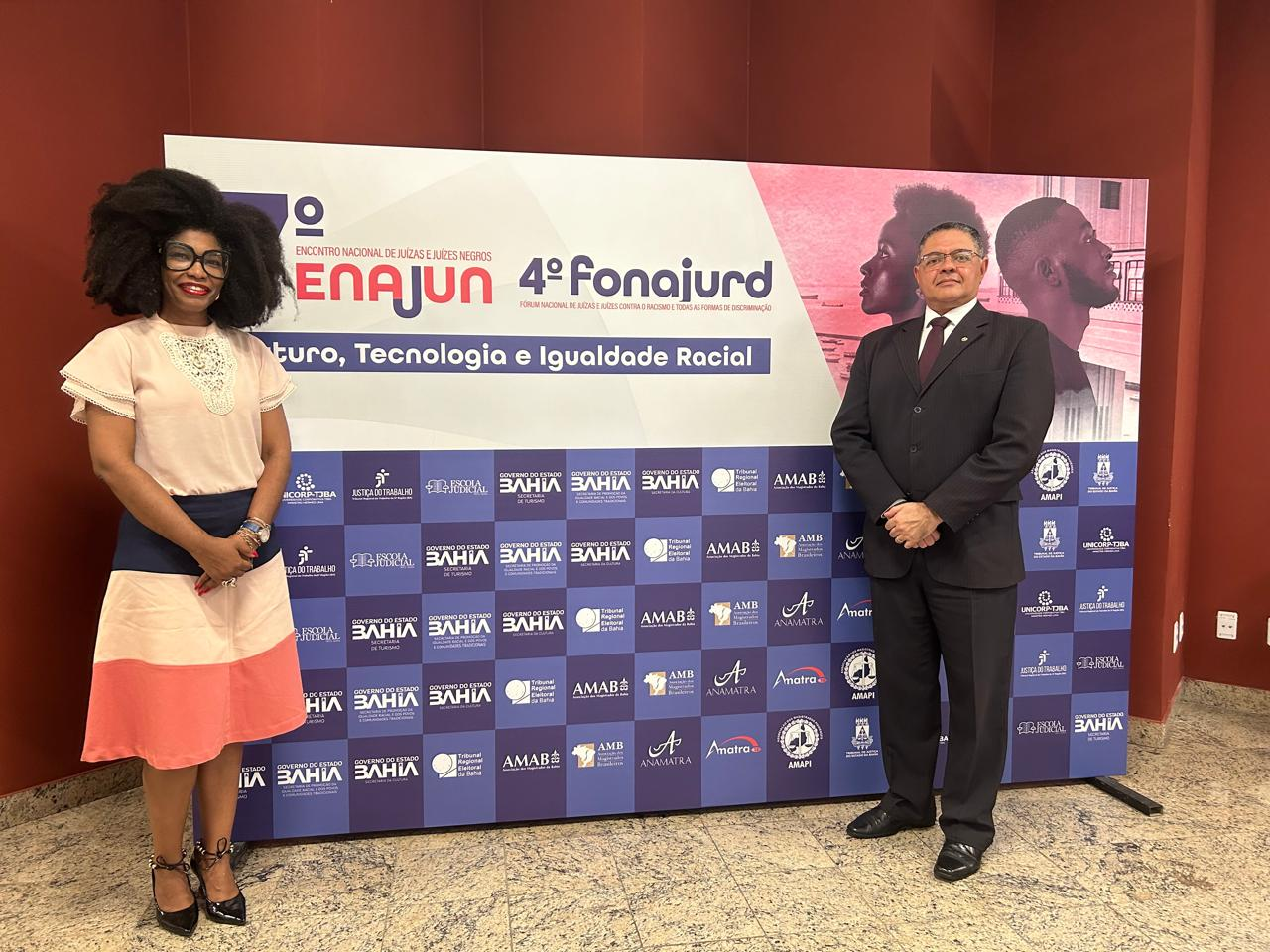 imagem mostra juíza Renata Nascimento e o juiz José Ribamar Mendes diante de banner com dizeres 7º Encontro Nacional de Juízas e Juízes Negros (ENAJUN) e 4º Fórum Nacional de Juízas e Juízes contra o Racismo e todas as formas de Discriminação (FONAJURD)