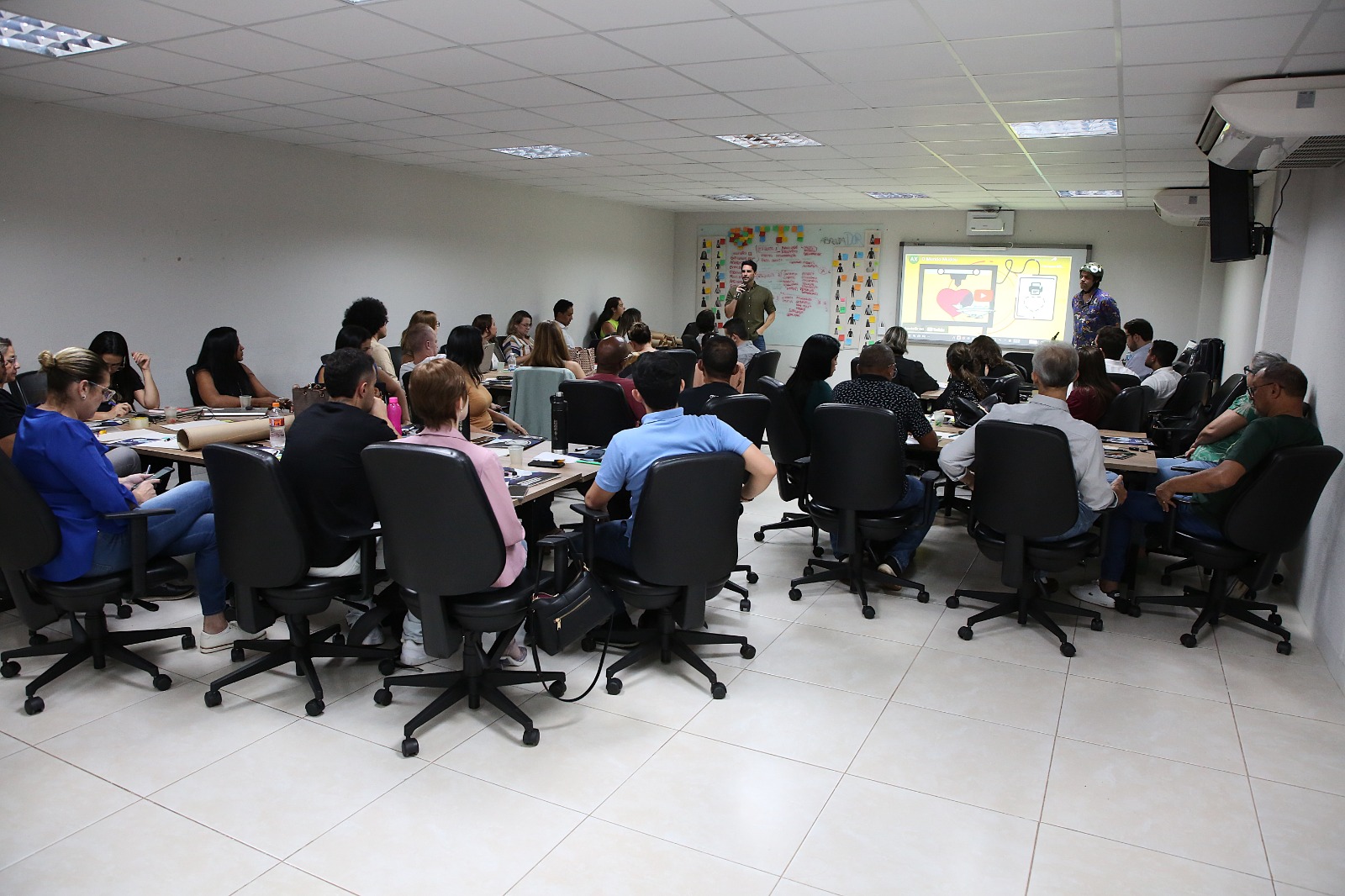 Sala de reunião com grupo de pessoas sentadas e concentradas. O piso é branco, paredes brancas e iluminação embutida no teto.