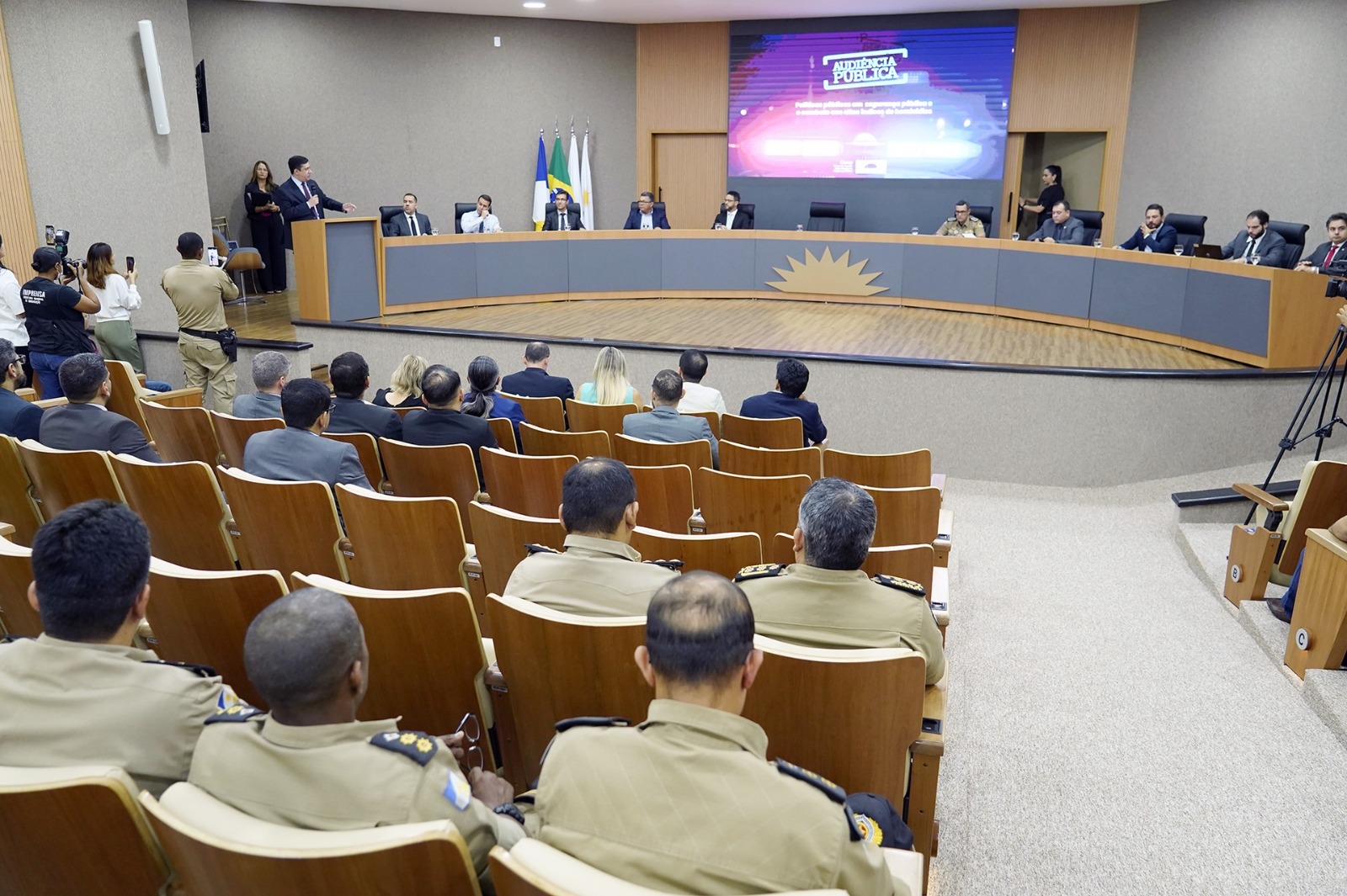 Imagem de um auditório, uma mesa grande ao fundo com pessoas sentadas e um telão no centro, ao fundo, e várias cadeiras e pessoas sentadas de costas