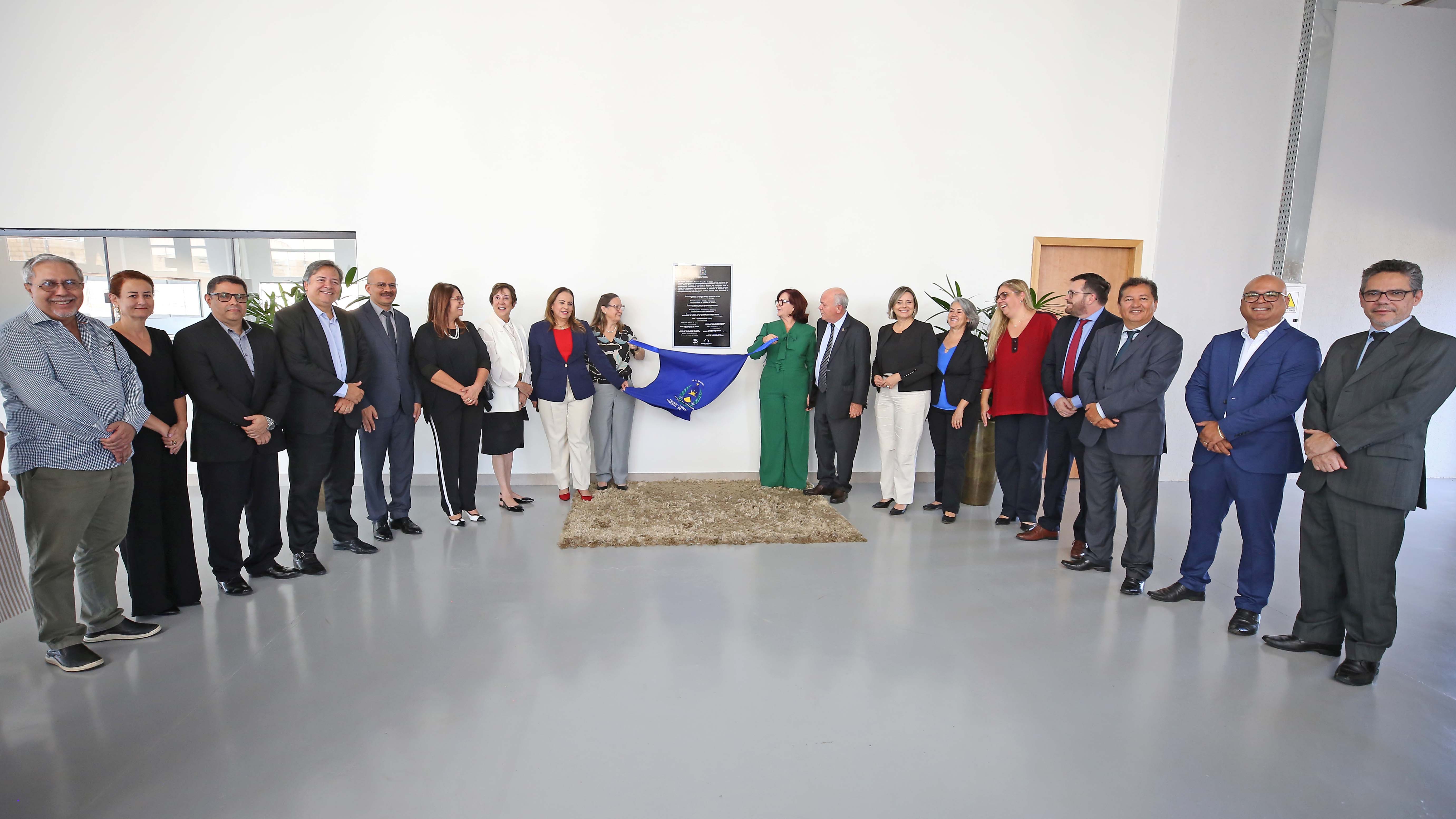 Homens e mulheres em pé, em frente a uma parede branca descerrando um tecido de uma placa durante inauguração de um prédio
