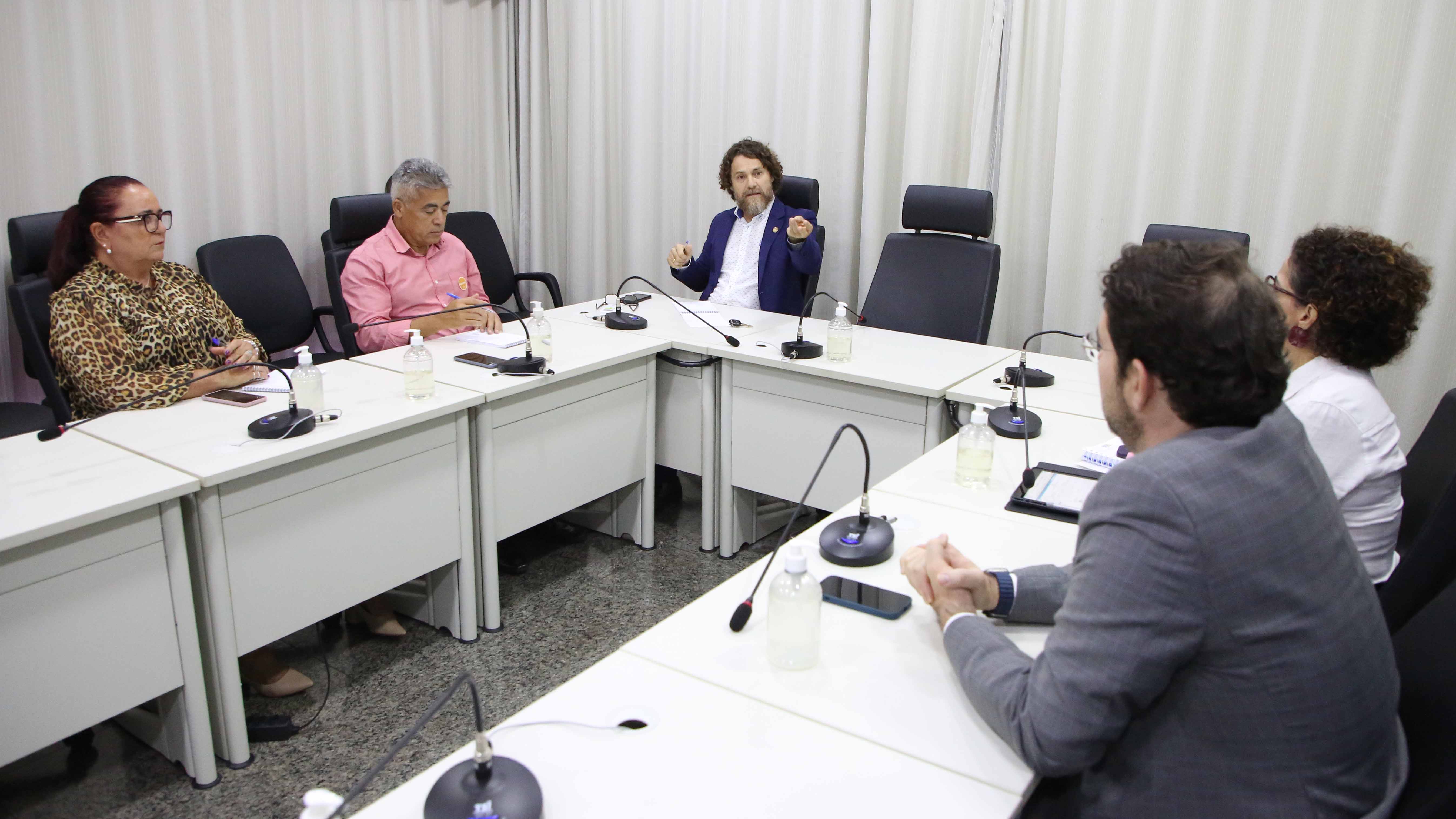 Juiz Océlio Nobre, de camisa branca e blazer azul, cabelos cacheados e barba, sentado à mesa ao fundo; ele participa de reunião e conversa com outras quatro pessoas sentadas: o juiz Roniclay Morais, o superintendente do Trabalho e Emprego, Jalson Jácomo; a coordenadora de Assessoramento da Diretoria-Geral do TJTO, Roberta Maciel; e a coordenadora da Ouvidoria, Alessandra Adorno
