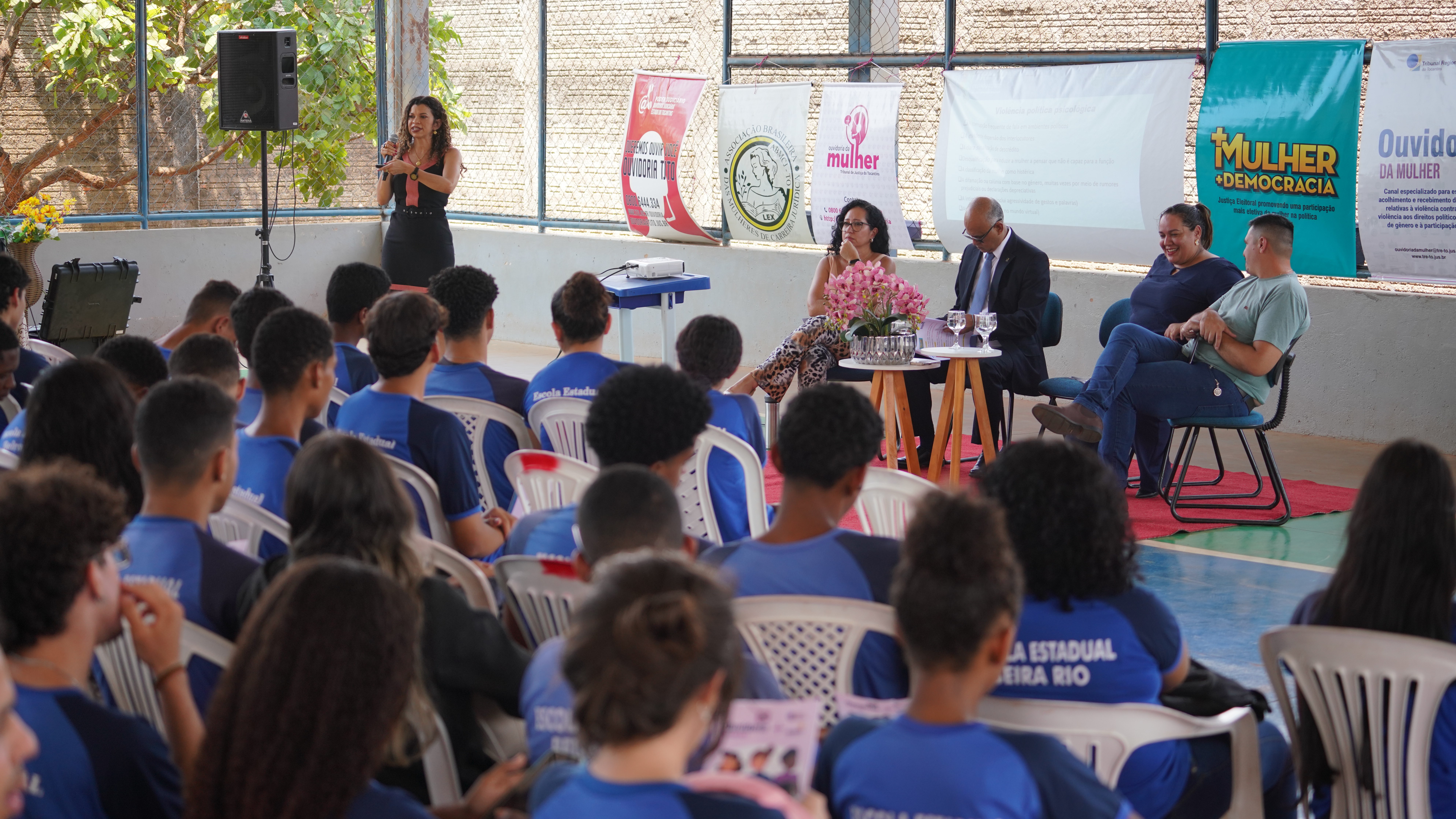 palestra, com vários alunos de costas sentados em cadeiras brancas, assistindo a palestra da juíza Edssandra, que está em pé à esquerda; ao centro, demais participantes da ação sentados