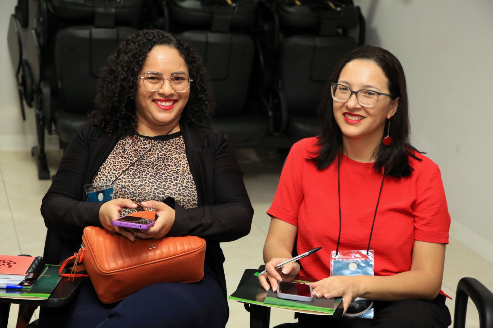 duas jovens senhoas sentadas, uma ao lado da outra; a primeira morena, cabelos cacheados, usando calça azul marinho, bluza estampada e blazer preto, segurando um celular e uma bolsa nas pernas; a outra da esquerda tem cabelos lisos pretos, usa óculos, blusa vermelha e calça preta