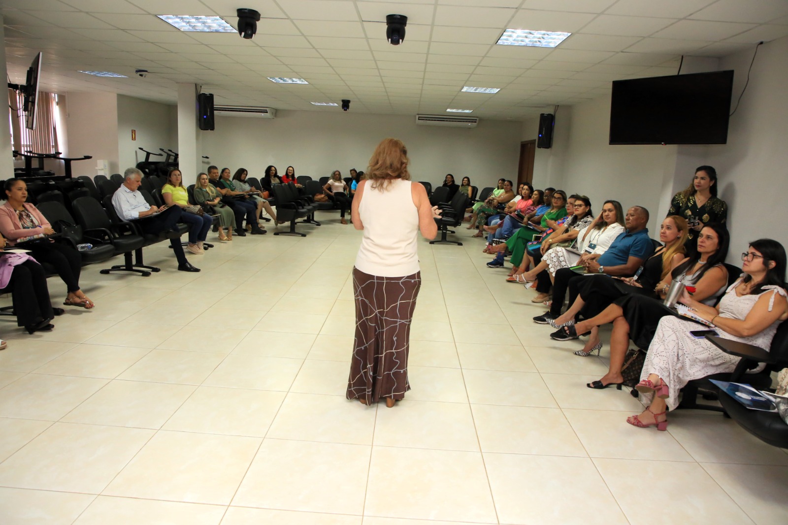 sala de aula com cadeiras em u com vários servidores sentados. De costas uma mulher, de cabelos louros, usando bluza branca e calça estampada nas cores marrom e branca,  fala aos servidores