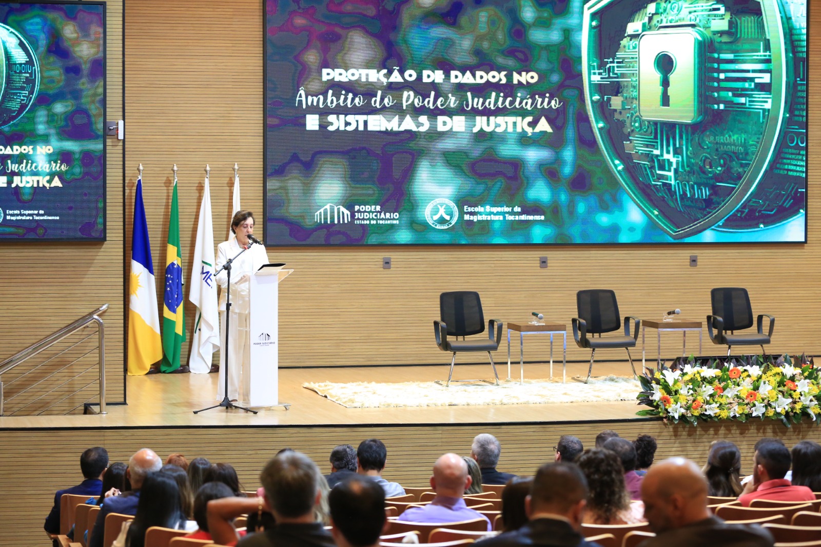 audiório com várias pessoas sentadas; no pauco a desembargadora Angela Haonat, uma mulher usando calça e blusa brancas, fala no pulpito localizado à esqueda; ao fundo três cadeiras escuras e um painel com a identidade visual do evento em tons de verde com uma fechadura 