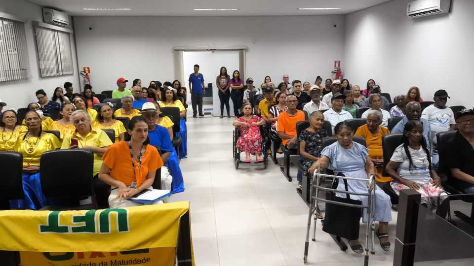 A imagem mostra um auditório com muitas cadeiras e fileiras de pessoas sentadas.  