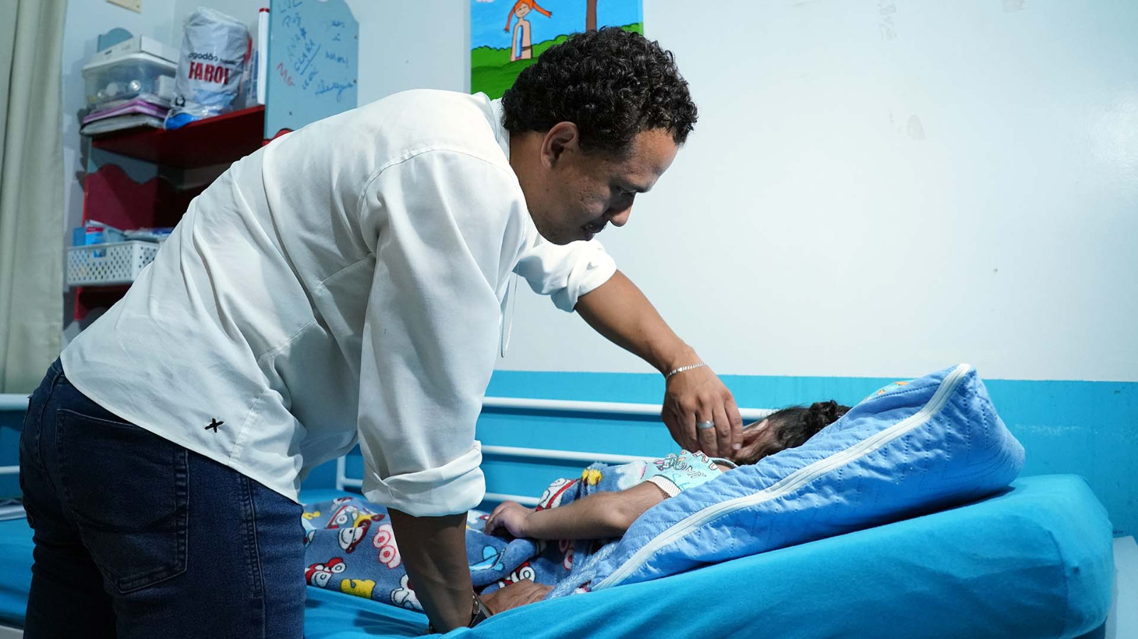 Marcelo, de camisa branca e calça presta, fazendo carinho em Lívia, que está deitada em uma cama, em seu quarto