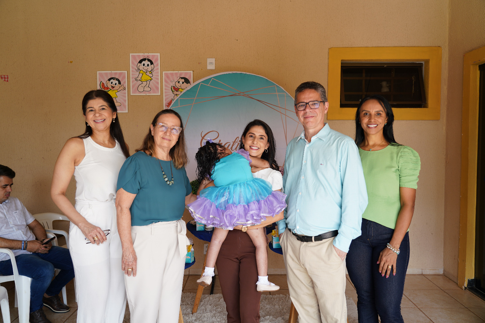 desembargadora maysa, de blusa verde e calça bege; juiz Adriano, de calça bege e camisa azul clara ao lado de duas servidoras e ao centro a madrinha da Lívia com a pequena nos braços, de costas e usando um vestido de tule nas coes azul e lilás