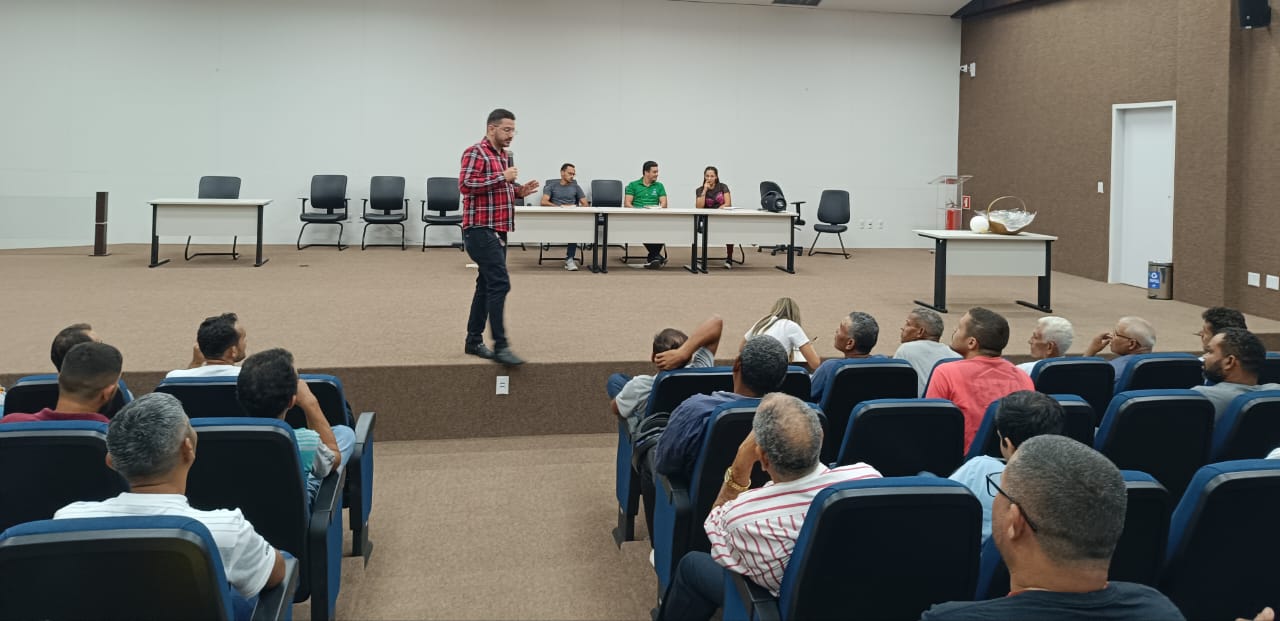 Auditório com paredes banca e marron e poltronas azuis. No palco o palestrante falando, um homem moreno claro de óculos e trajando uma camisa xadrez em tons vermelho, branco e preto e calça preta. Nas cadeiras, vários reeducandos sentados de costas assistindo a palestra
