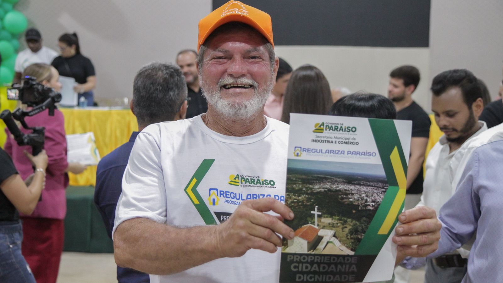 Senhor com camiseta branca, barba e boné laranja posando para foto com um sorriso no rosto segurando um documento.