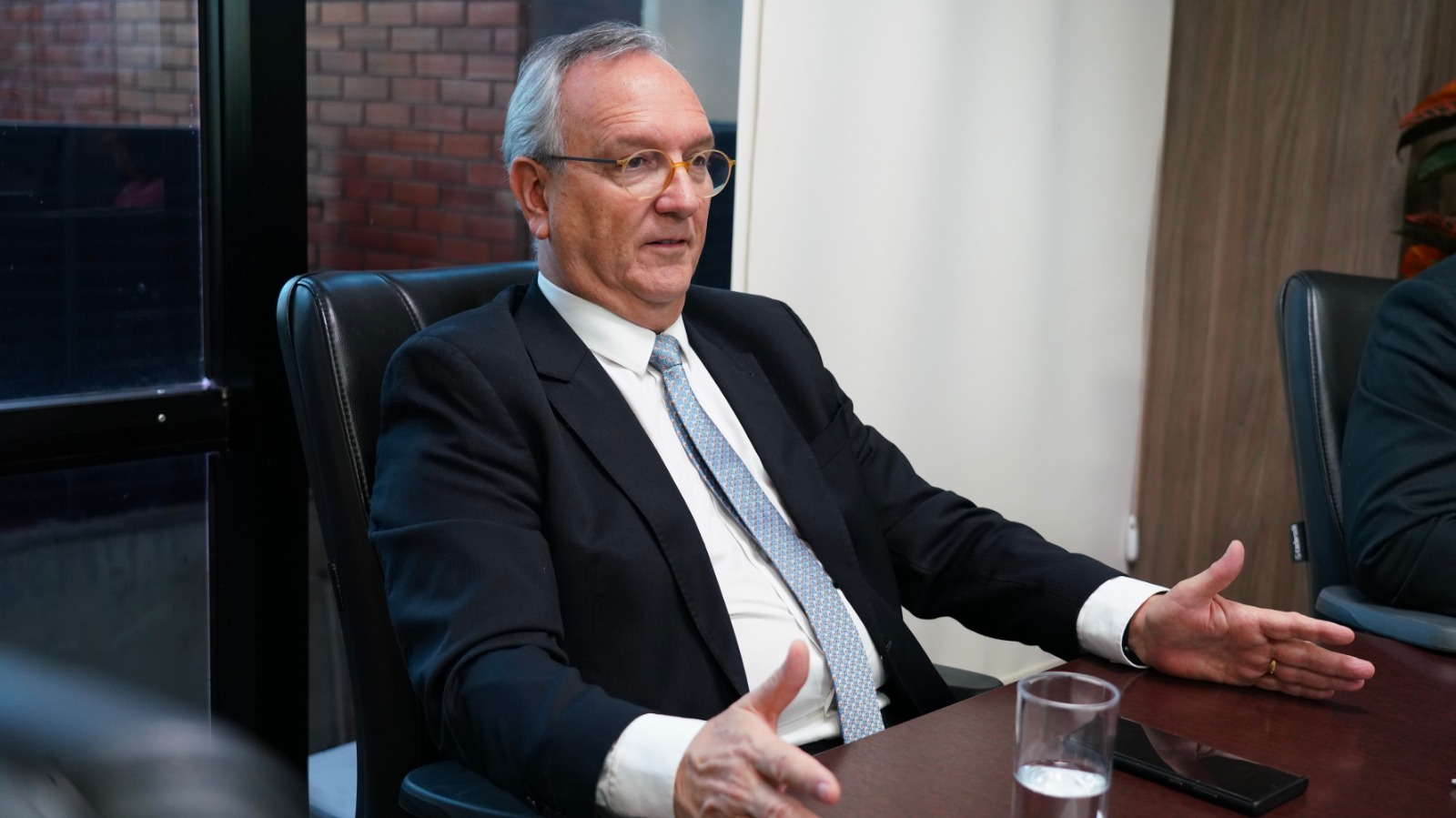 desembargador Pedro Nelson sentado, em seu gabinete; usa terno preto, camisa branca e gratava azul claro. Homem branco, cabelos grisalhos, usa óculos