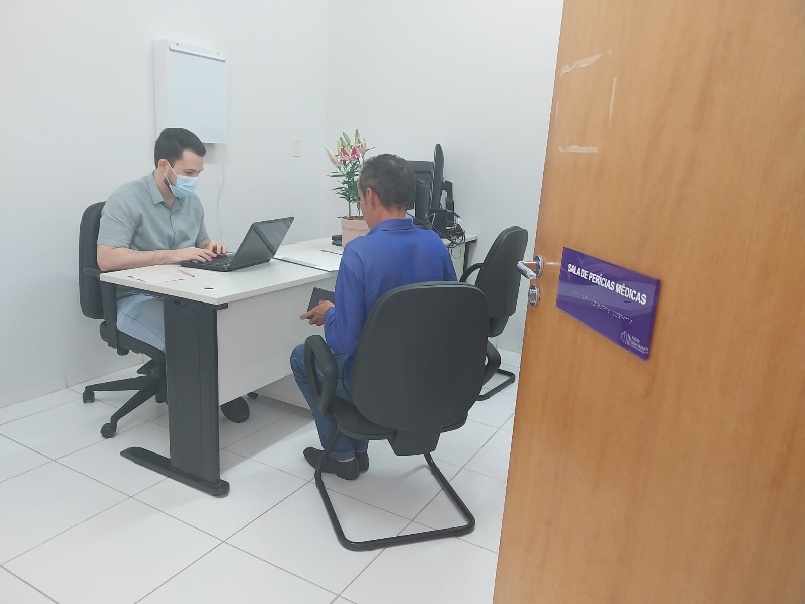 médico atendendo um paciente para perícia médica previdenciária; o médico está sentado, usando um notebook e o paciente, um homem, está sentado de costa; em primeiro plano uma porta aberta de madeira com uma placa azul escrita Sala de Perícias Médicas em fonte branca