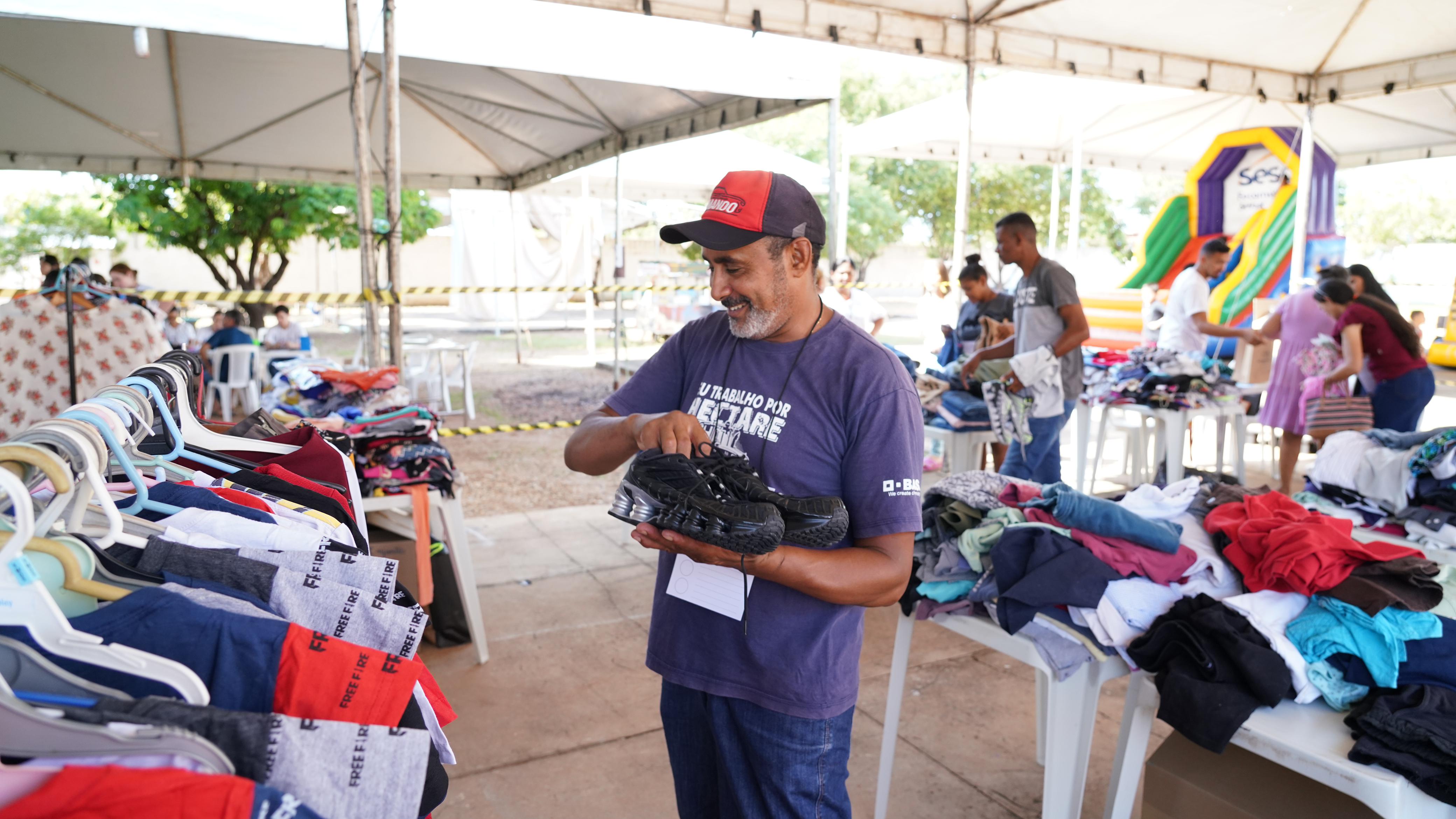 Homem de boné vermelho e preto e camiseta roxa segura um par de sapatos no Varal Solidário