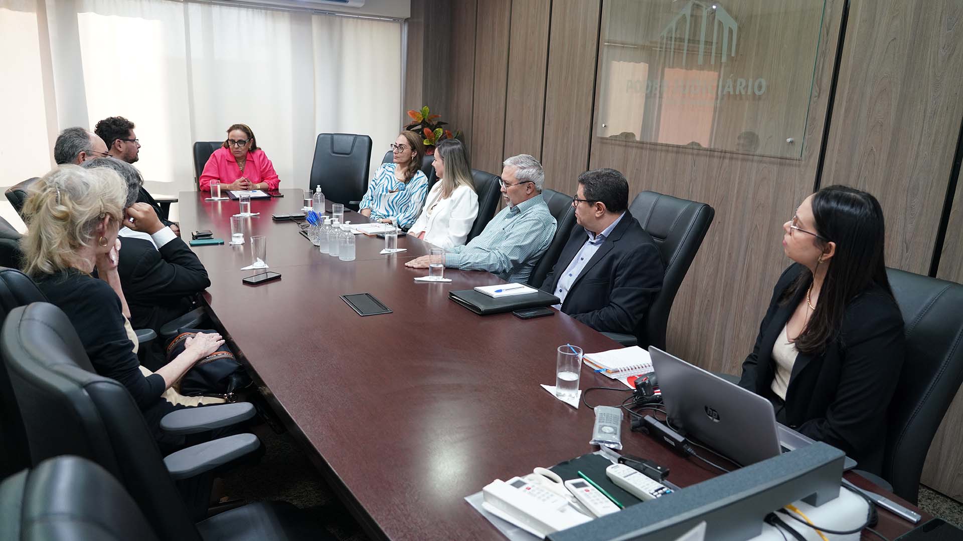 Em uma sala com uma mesa de reuniões, parede de madeira, janelão de vidro com uma cortina branca; na cabeceira da mesa, a desembargadora Ângela Prudente, de roupa rosa e óculo, preside a reunião do grupo de trabalho, formado po magistrados e diretores do TJTO 