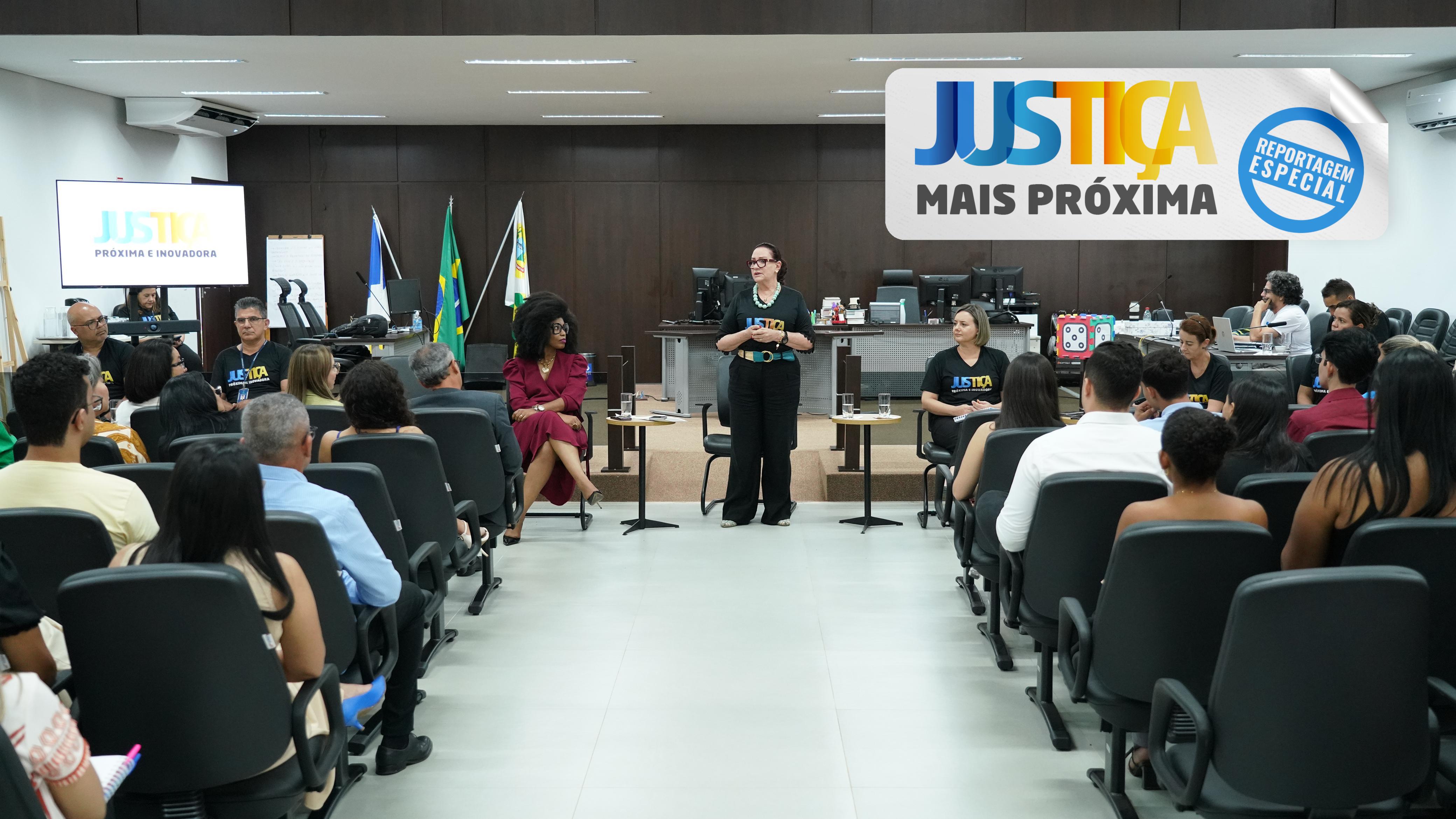 A imagem mostra uma sala onde várias pessoas estão sentadas em cadeiras pretas. Elas olham para duas mulheres que estão em pé na frente, falando. Uma mulher veste um vestido escuro vinho e a outra usa uma camiseta preta. Atrás delas, há uma tela com o logo "Justiça Próxima e Inovadora" e também bandeiras do Brasil. A sala é bem iluminada.