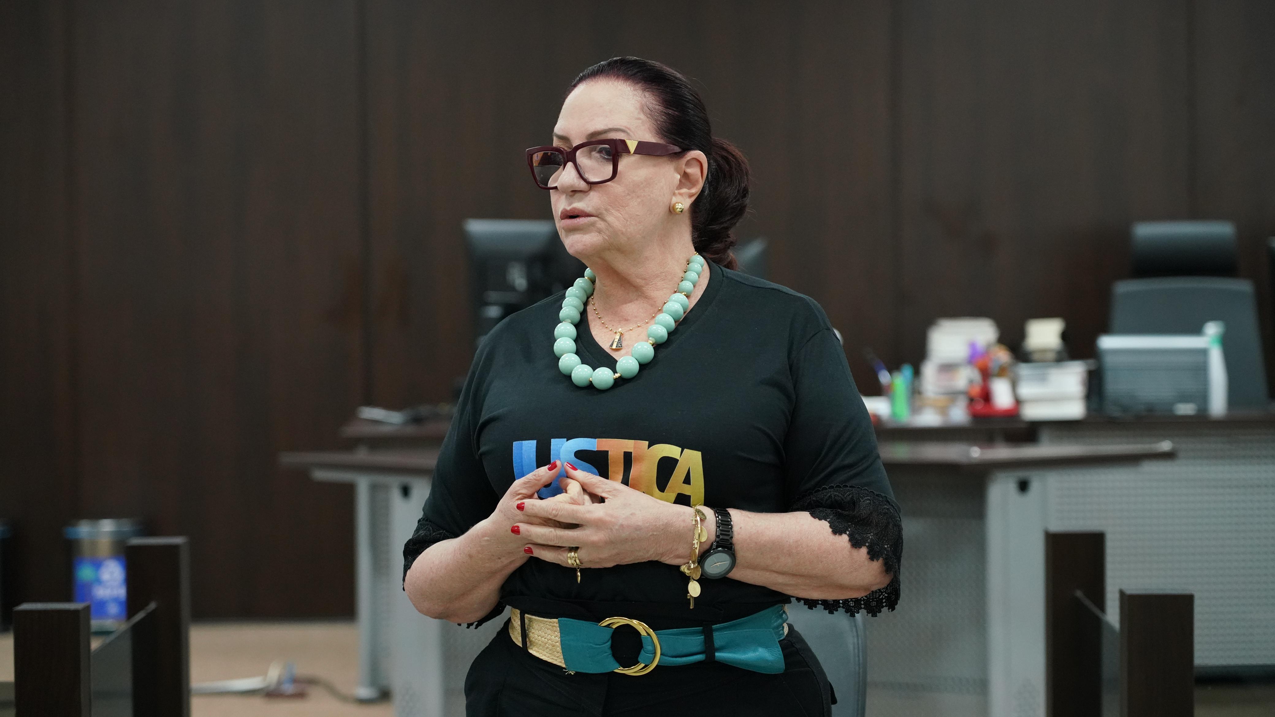 A imagem mostra uma mulher de pele clara, com cabelos escuros presos num coque baixo. Ela usa óculos de armação escura, brincos pequenos e dourados e um colar de contas verde-azulado. Veste uma camiseta preta com a palavra "JUSTIÇA" escrita nela em letras coloridas e um cinto largo com uma fivela dourada e uma faixa de tecido verde-azulado. Ela tem as mãos juntas na altura do peito e parece estar falando ou prestes a falar. O fundo é desfocado, mas mostra um ambiente de escritório com uma mesa e cadeiras, sugerindo que ela possa estar numa reunião ou apresentação. A mulher aparenta ser séria e concentrada.